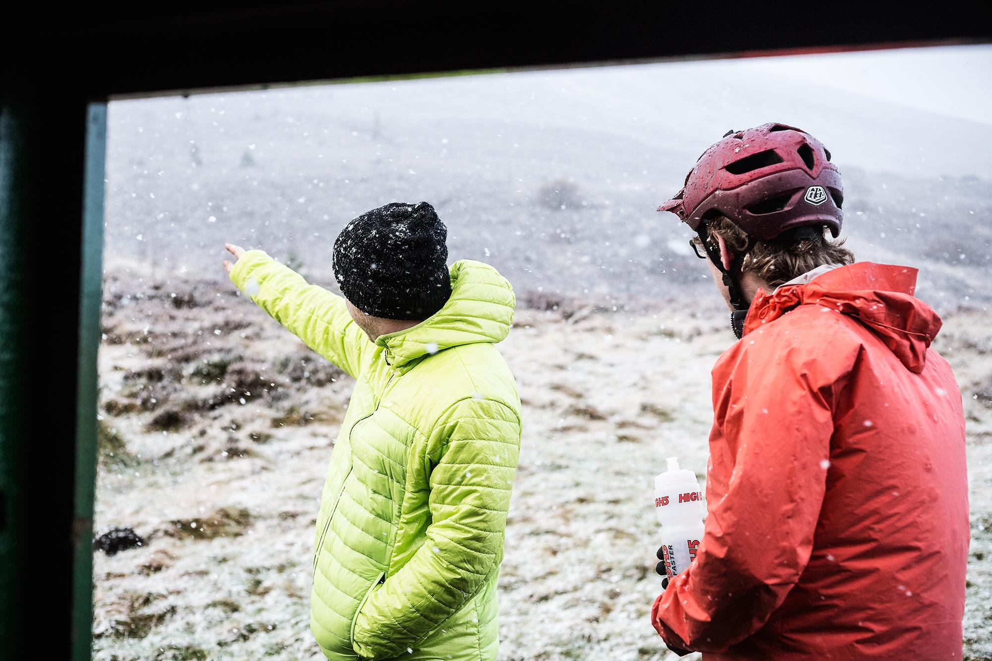 Two individuals are standing outside in a snowy environment. One person, wearing a bright green jacket and a black beanie, is pointing towards something in the distance, while the other, dressed in a red jacket and wearing a cycling helmet, is holding a water bottle. The landscape appears cold and snowy, indicating winter conditions. Rhyvoan Pass mountain bike trail.