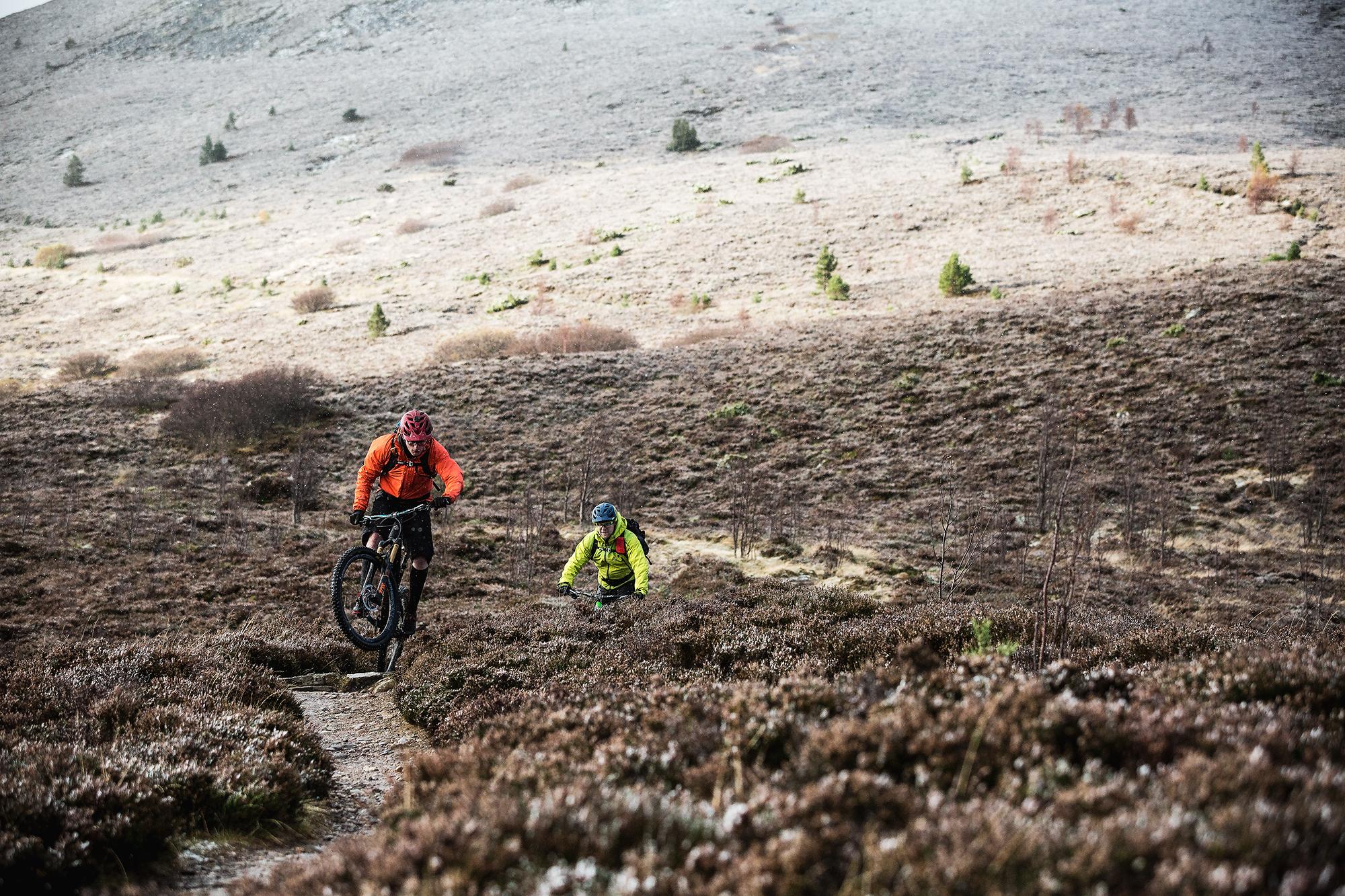 Two mountain bikers navigating a rugged terrain. One rider in an orange jacket performs a wheelie on a rocky path, while the other in a yellow jacket climbs steadily uphill. The landscape features sparse vegetation and a hilly backdrop under a cloudy sky. Meall a Bhuachaille Trail mountain bike trail.