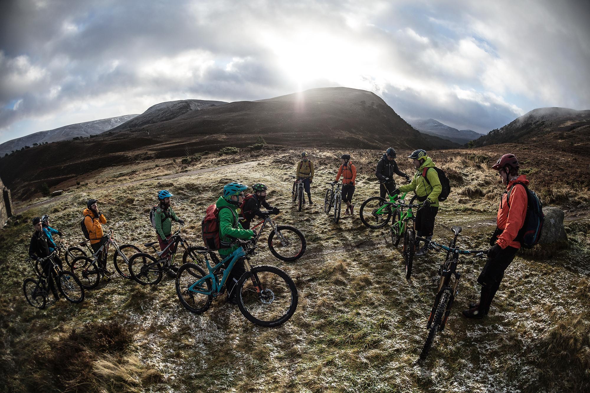 A group of mountain bikers gathered on a grassy area surrounded by hills. They are wearing colorful jackets and helmets, with some bicycles leaning against them. The scene is illuminated by sunlight peeking through clouds in the background. Some snow can be seen on the ground, suggesting a cooler climate. Meall a Bhuachaille Trail mountain bike trail.