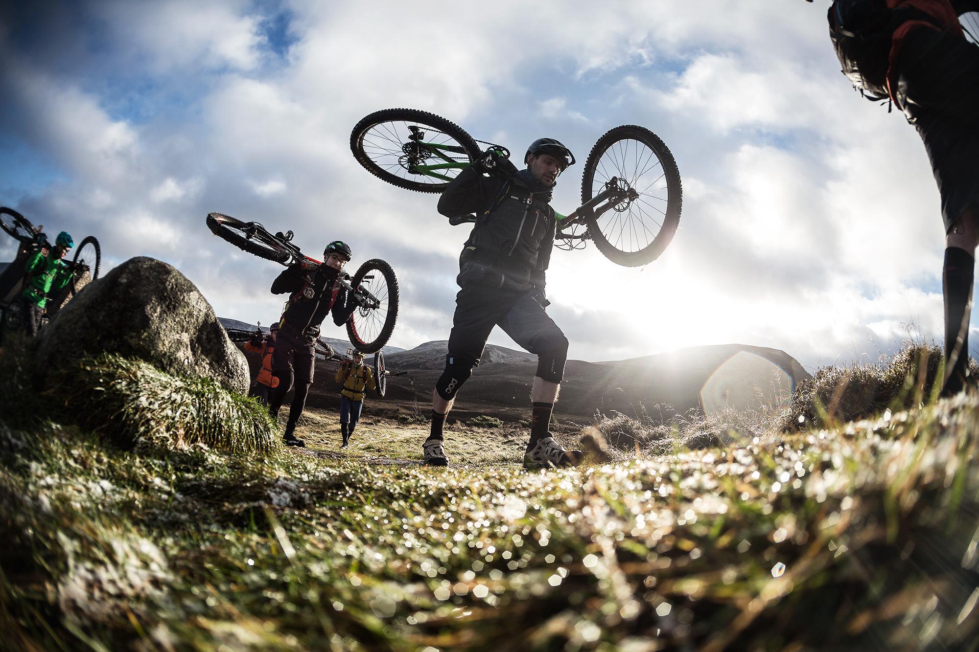 A group of mountain bikers carrying their bikes over a grassy landscape, with a dramatic sky and sunlight shining in the background. The riders are dressed in outdoor gear, focusing on navigating the terrain as they trek uphill. Meall a Bhuachaille Trail mountain bike trail.