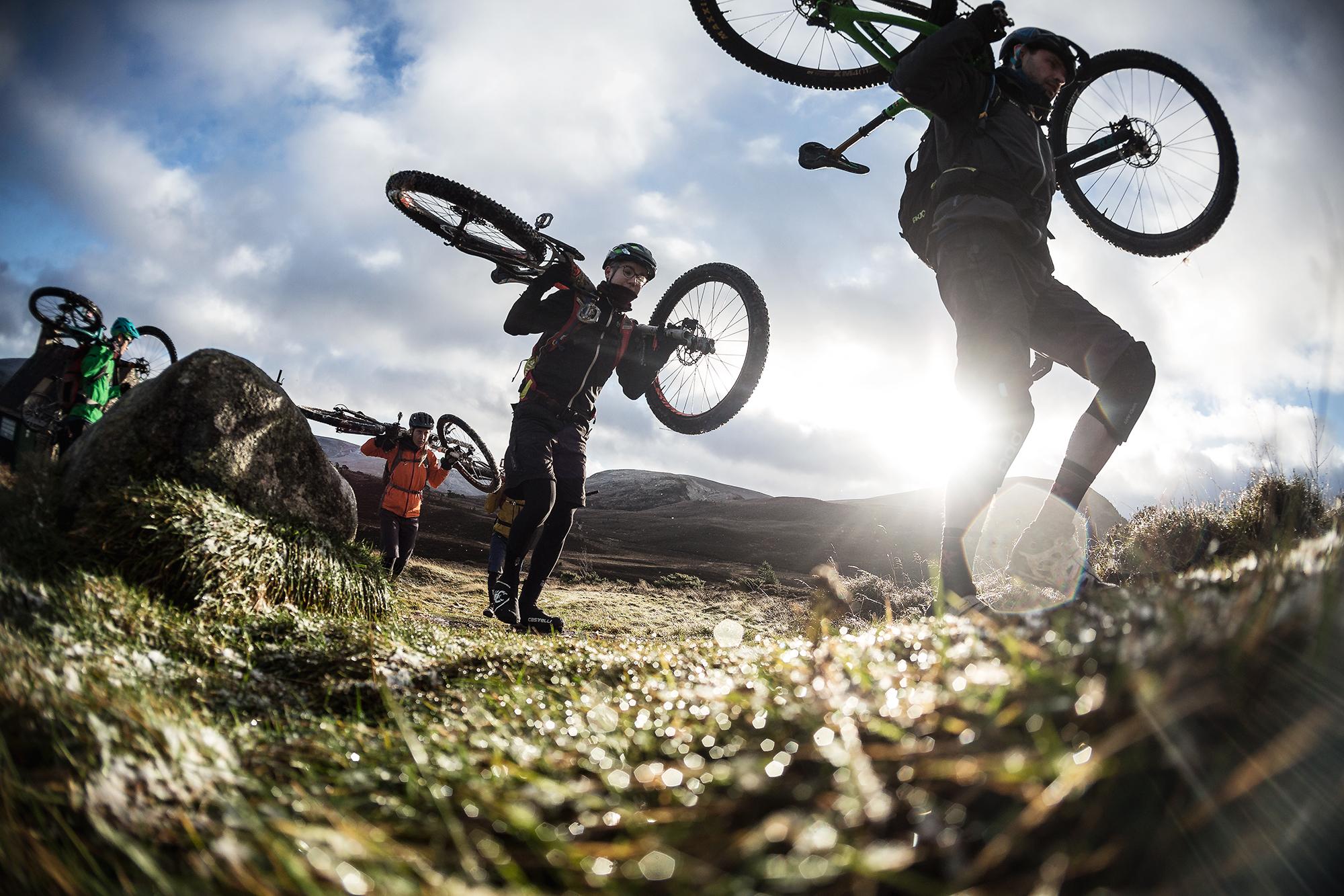 A group of mountain bikers carrying their bikes on a grassy trail, with sunlight shining in the background and a cloudy sky above. The riders are dressed in various outdoor gear, showcasing a vibrant mix of colors. The scene conveys a sense of adventure and the challenges of biking in rugged landscapes. Meall a Bhuachaille Trail mountain bike trail.