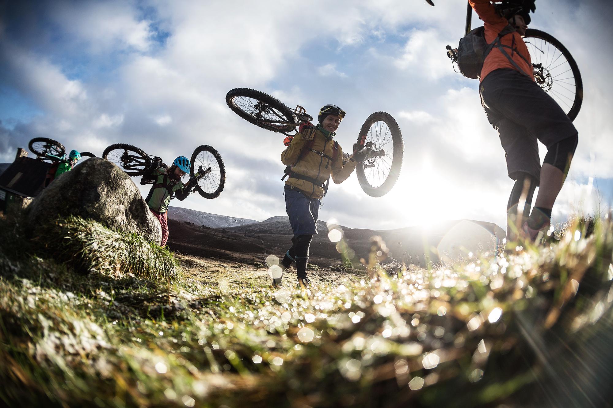 A group of mountain bikers carrying their bikes over rocky terrain, with sunlight shining through clouds in the background. The riders wear helmets and outdoor clothing, showcasing their active lifestyle in a scenic, rugged landscape. Meall a Bhuachaille Trail mountain bike trail.