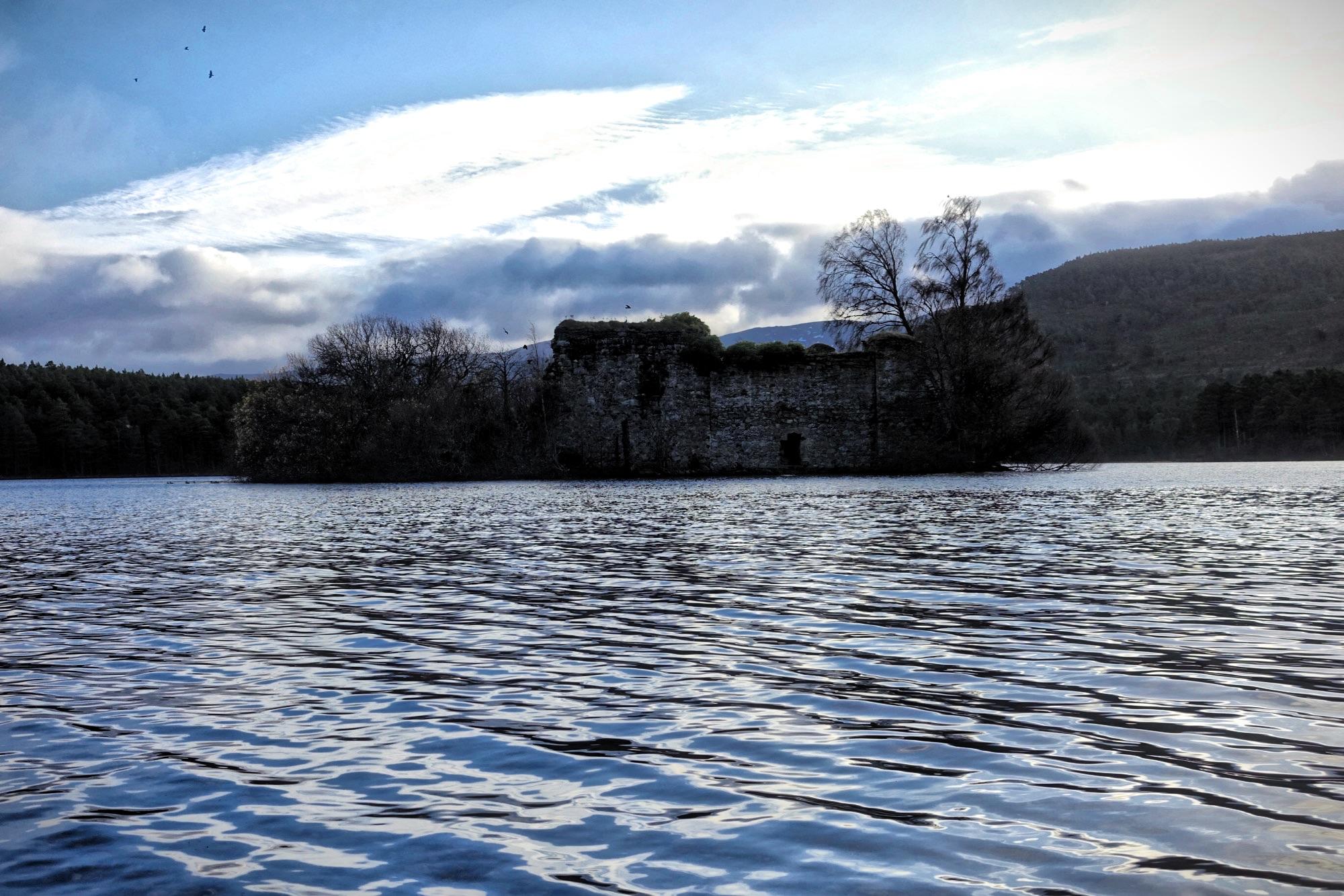 A serene lakeside view featuring a partially submerged stone ruin surrounded by trees. The water reflects the cloudy sky, with gentle ripples suggestive of a calm atmosphere. Birds can be seen flying above, while mountains rise in the background, completing the tranquil landscape. Loch an Eilein mountain bike trail.