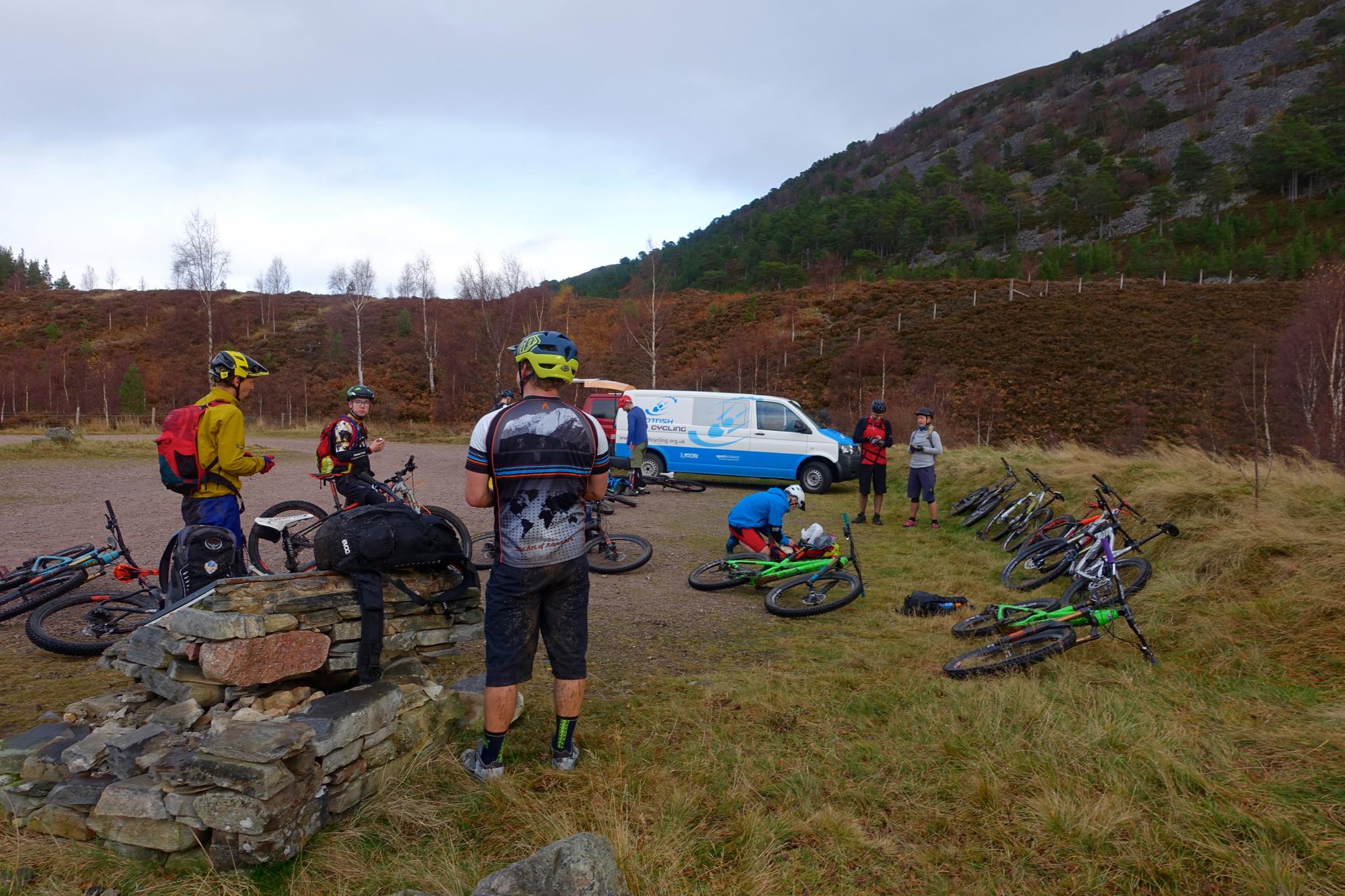 A group of mountain bikers gathered at a stop in a natural setting, with several bicycles parked nearby. Some riders are interacting with each other while others are inspecting or adjusting their bikes. A van is visible in the background, and the landscape features rolling hills and sparse vegetation. River Feshie Trail mountain bike trail.