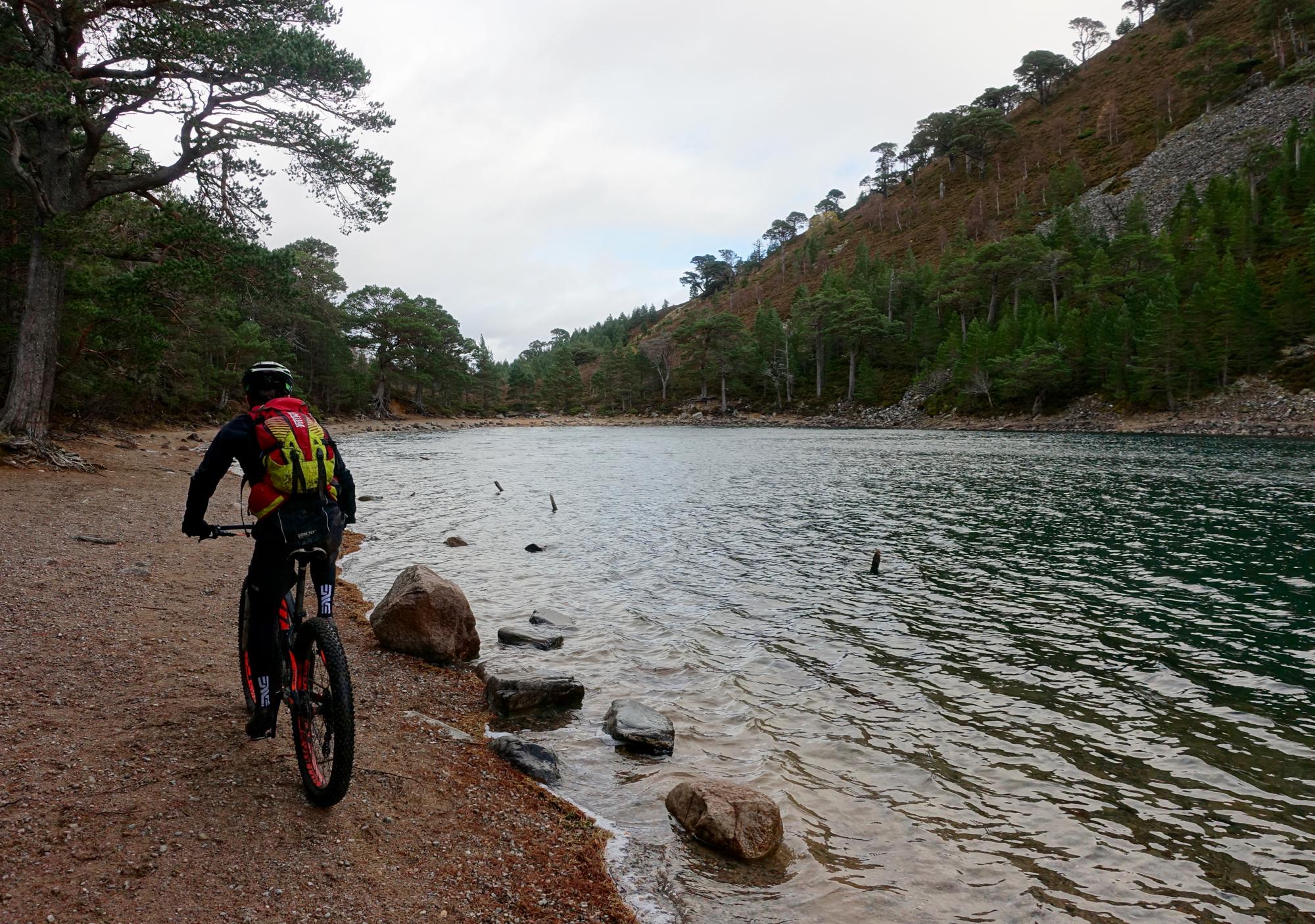 A cyclist with a backpack rides along a gravel path beside a calm lake, surrounded by lush green trees and rocky hills under a cloudy sky. Rhyvoan Pass mountain bike trail.