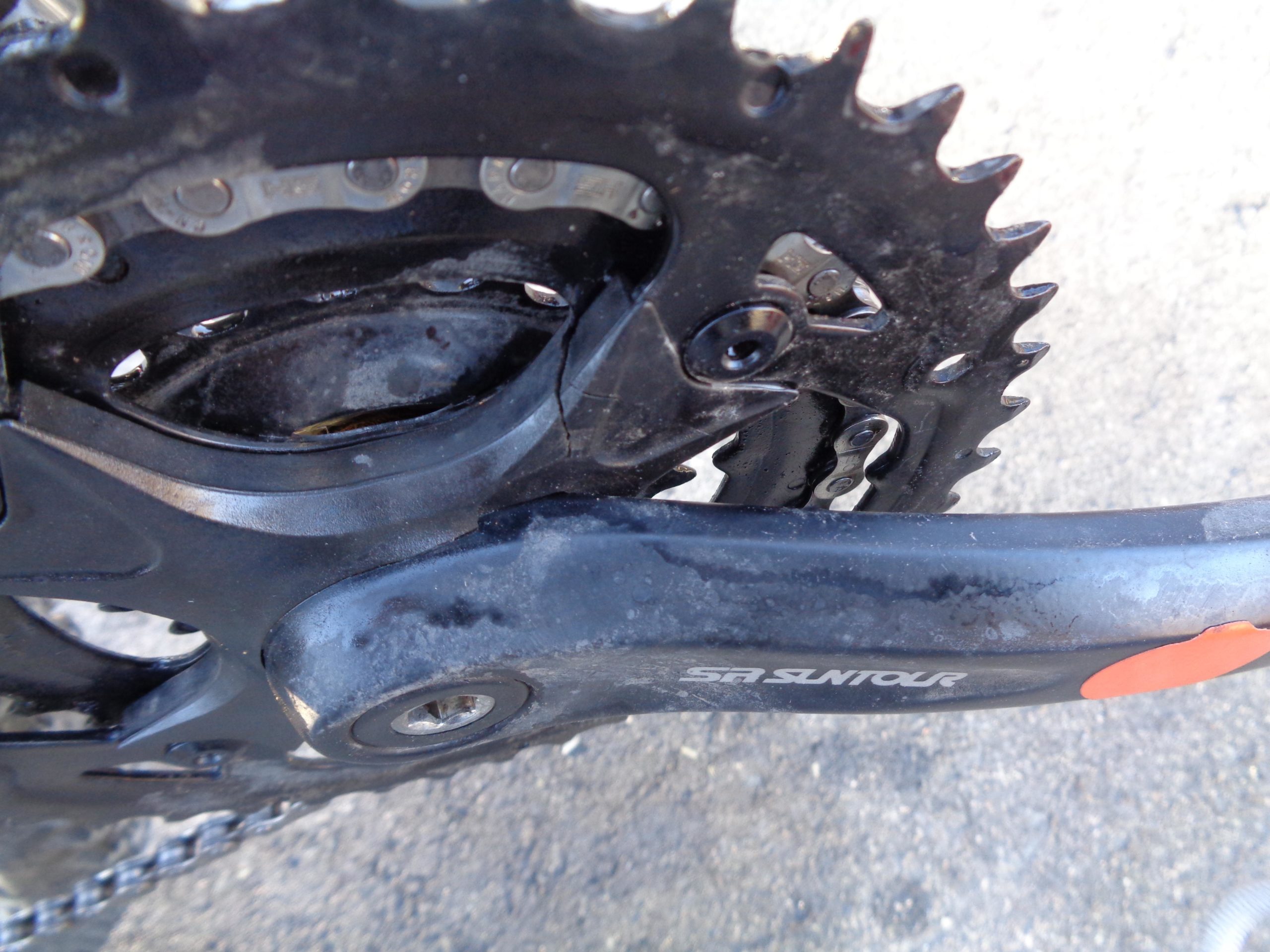 Close-up image of a bike's crankset and chainring, showing the gears and chain in detail. The components are slightly worn, highlighting signs of use. The crank arm has a logo that reads "SR SUNTOUR." The background is a textured surface, adding context to the mechanical focus of the image. Wild Turkey mountain bike trail.