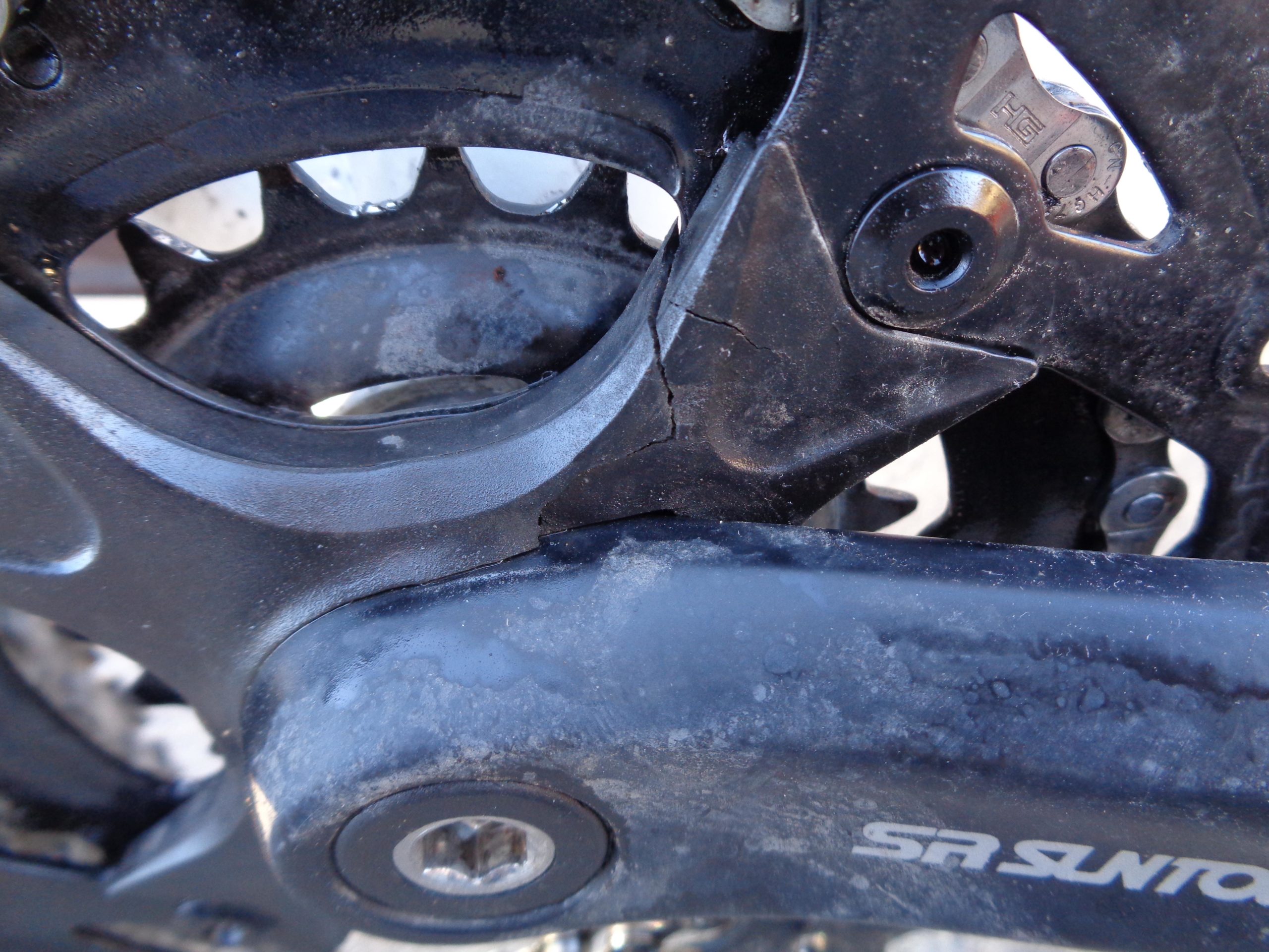 Close-up image of a bicycle crankset showing wear and damage. The metal components are black with visible scratches and signs of use. A noticeable crack is present near the connection point of the crank arm, indicating potential issues with the bike's drivetrain. Wild Turkey mountain bike trail.