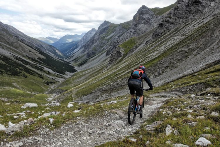 A mountain biker navigating a rocky trail in a steep valley, surrounded by towering mountains and lush greenery under a cloudy sky.
