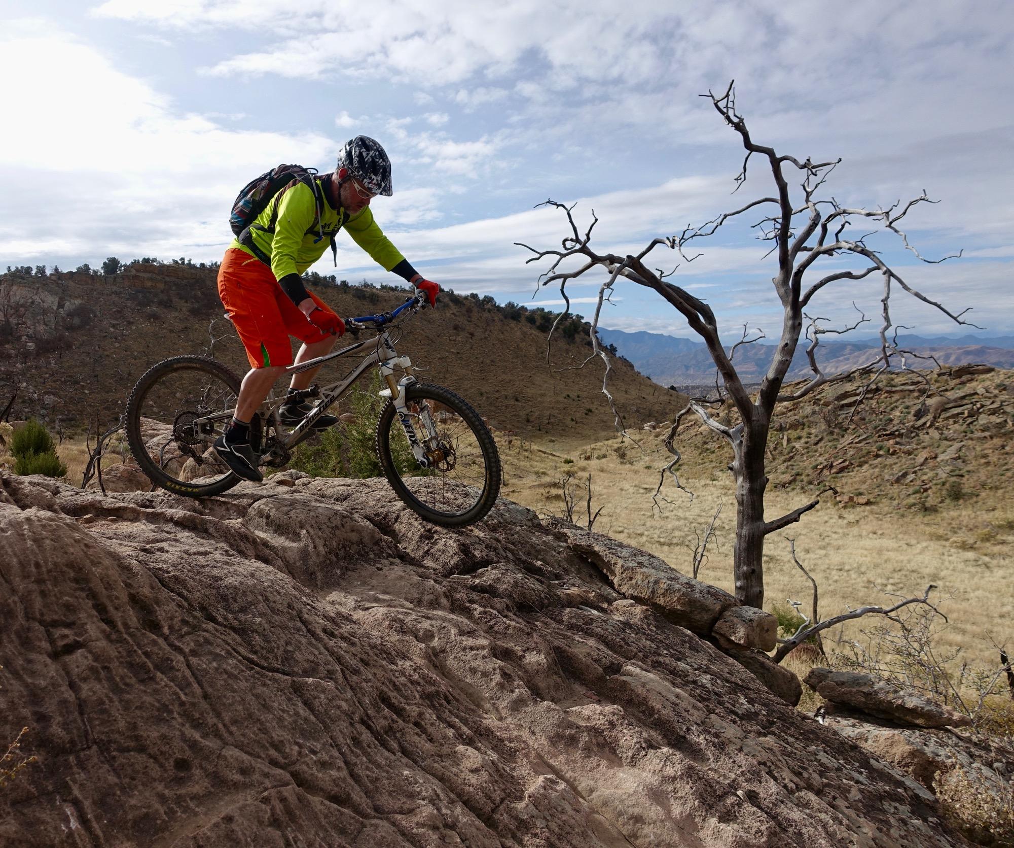 A mountain biker wearing a bright green long-sleeve shirt and orange shorts navigates a rocky terrain. In the background, a barren, twisted tree stands against a rugged landscape of hills and sparse vegetation under a cloudy sky. Oil Well Flats mountain bike trail.