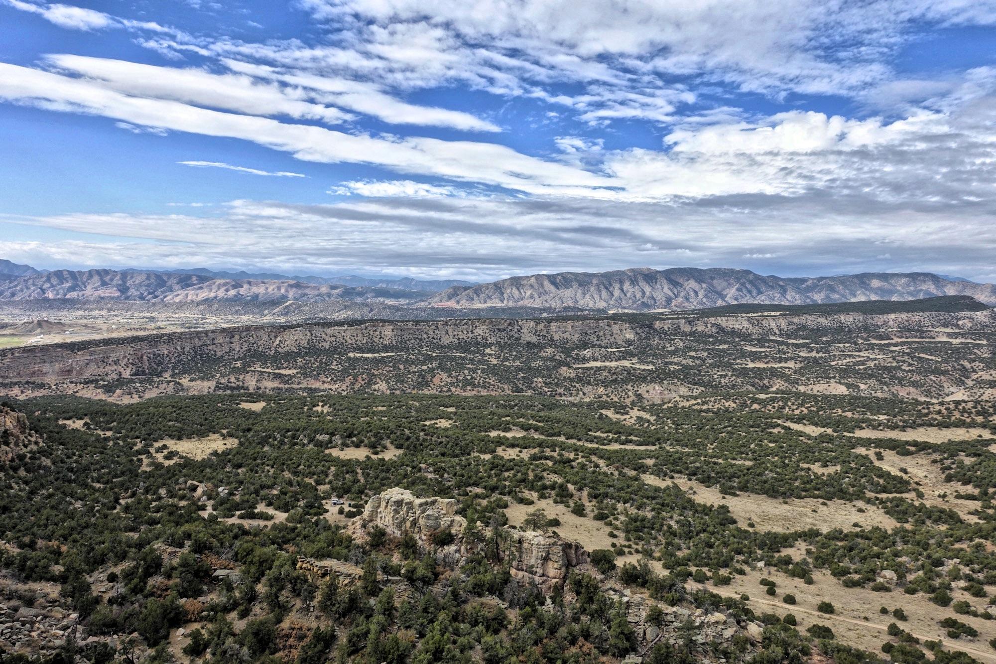 Panoramic view of a mountainous landscape with rolling hills and scattered greenery under a partly cloudy sky. The foreground features a rocky outcrop, while the distant mountains create a dramatic backdrop. The image captures the natural beauty and vastness of the area. Oil Well Flats mountain bike trail.