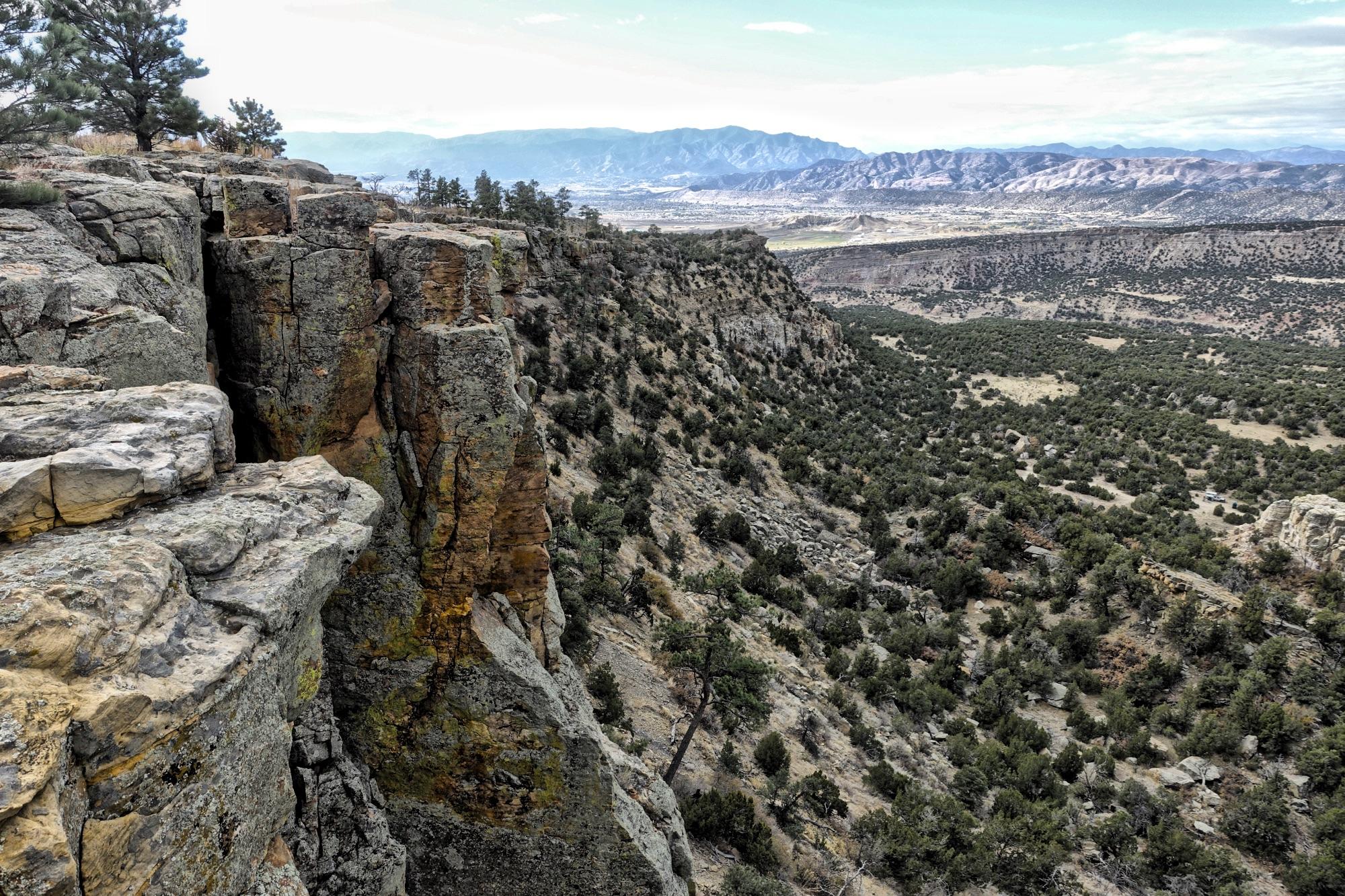 A rocky cliff edge overlooking a vast landscape of rolling hills, greenery, and distant mountains under a cloudy sky. The foreground features jagged rock formations, while the background extends into a valley filled with trees and shrubs, showcasing a rugged and natural terrain. Oil Well Flats mountain bike trail.