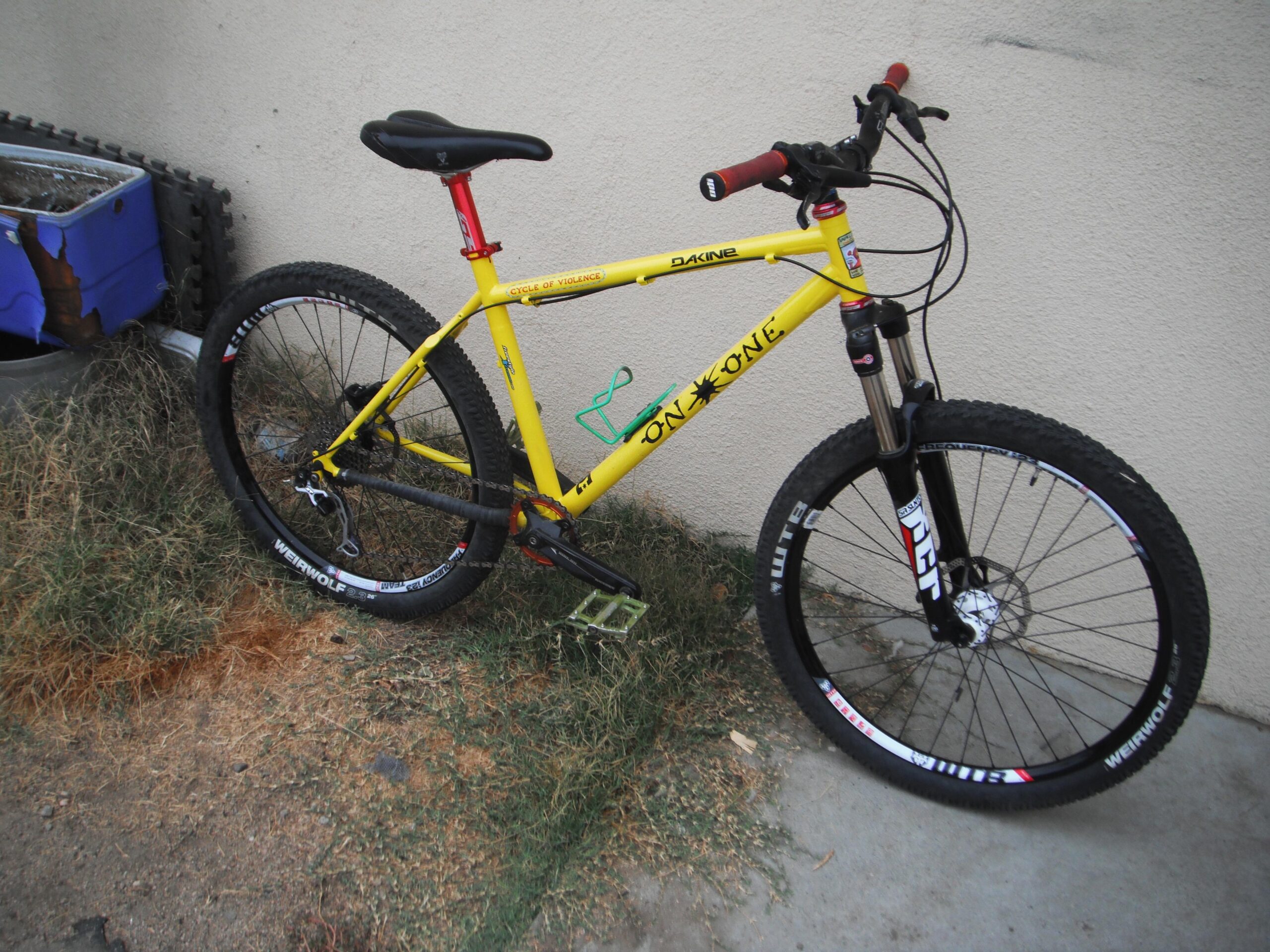 On-One 456 Evo2: A bright yellow mountain bike with thick tires, featuring red handlebars and a black front suspension fork. The bike is leaning against a wall, positioned on uneven ground with patches of grass and dirt around it. In the background, there is a blue container partially visible.