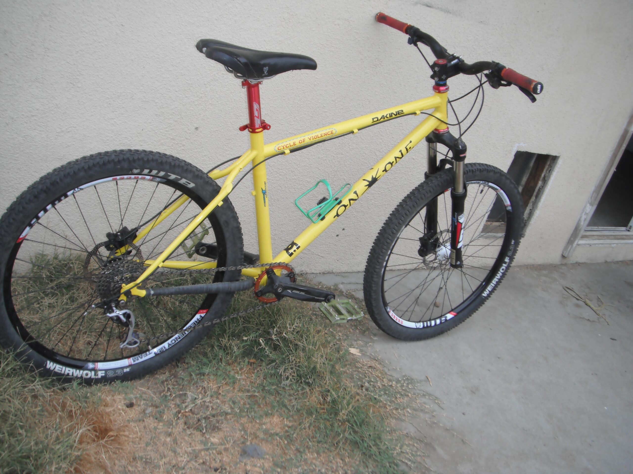 On-One 456 Evo2: A yellow mountain bike leaning against a wall with a grassy area in the foreground. The bike features large, knobby tires and a sturdy frame, with visible branding and components. A door is partially visible in the background.