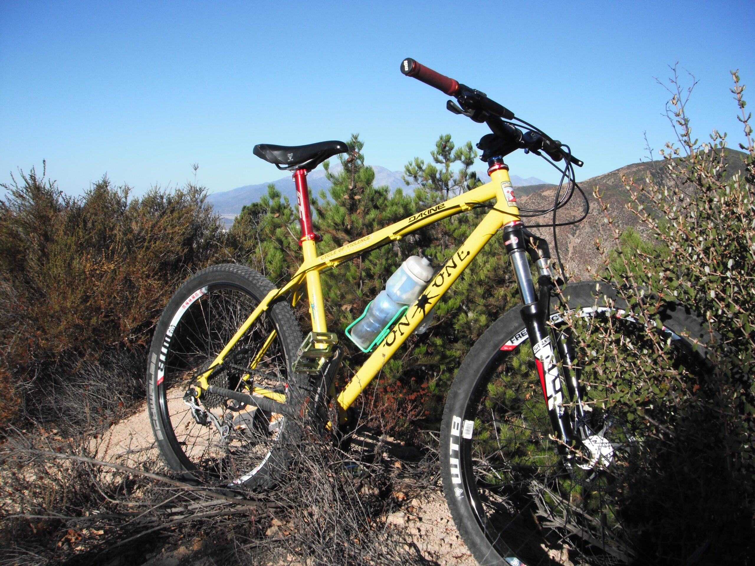 On-One 456 Evo2: A yellow mountain bike parked on rocky terrain surrounded by shrubs and small trees, with a blue water bottle attached to the frame. The clear blue sky and distant hills are visible in the background.