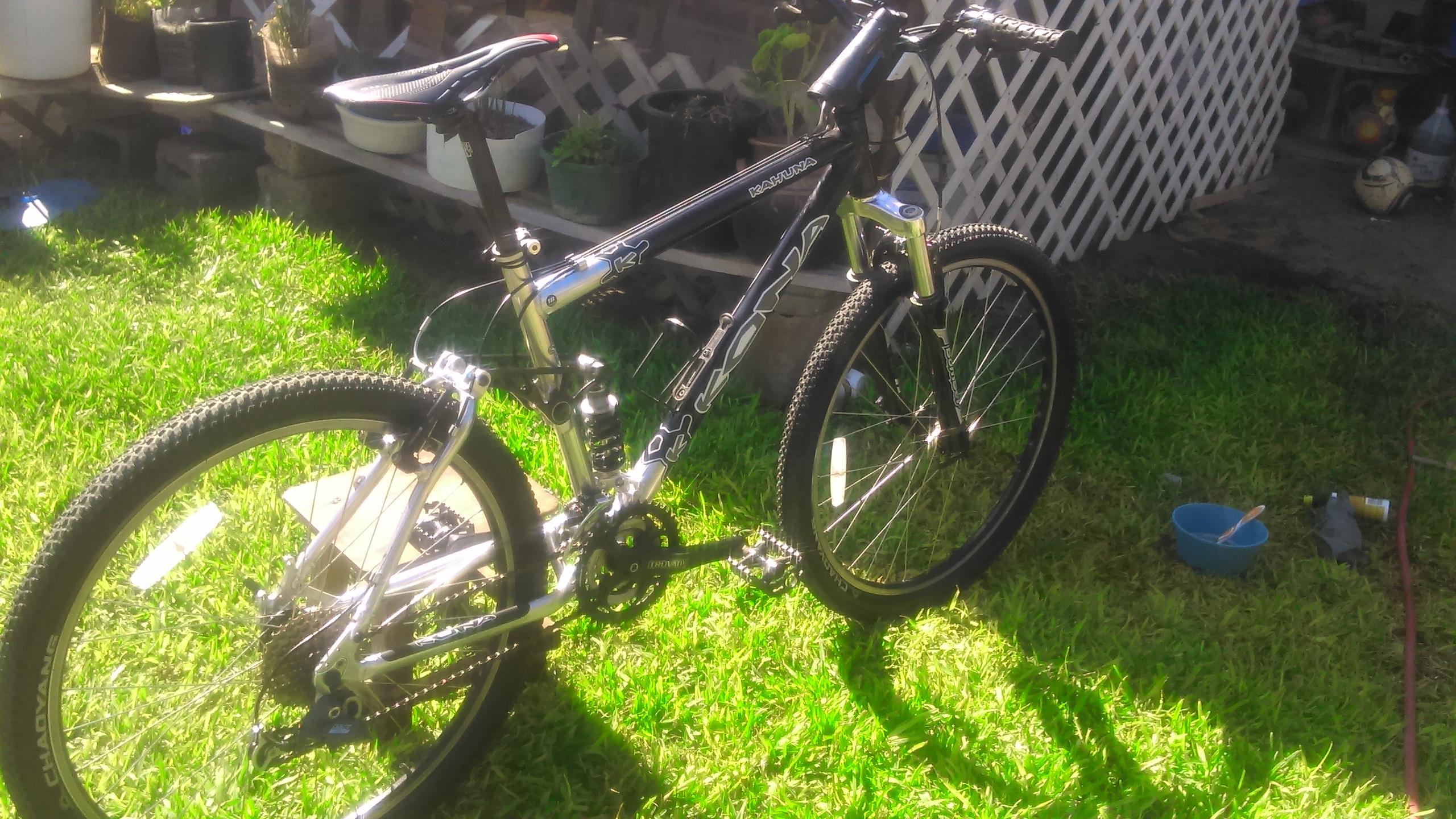 Kona Kahuna: A silver and black mountain bike is resting on green grass, with a clear blue sky in the background. The bike features thick, textured tires and a sturdy frame. It is positioned near a wooden structure with potted plants, and there are tools and a small bowl nearby, suggesting maintenance or repair work.