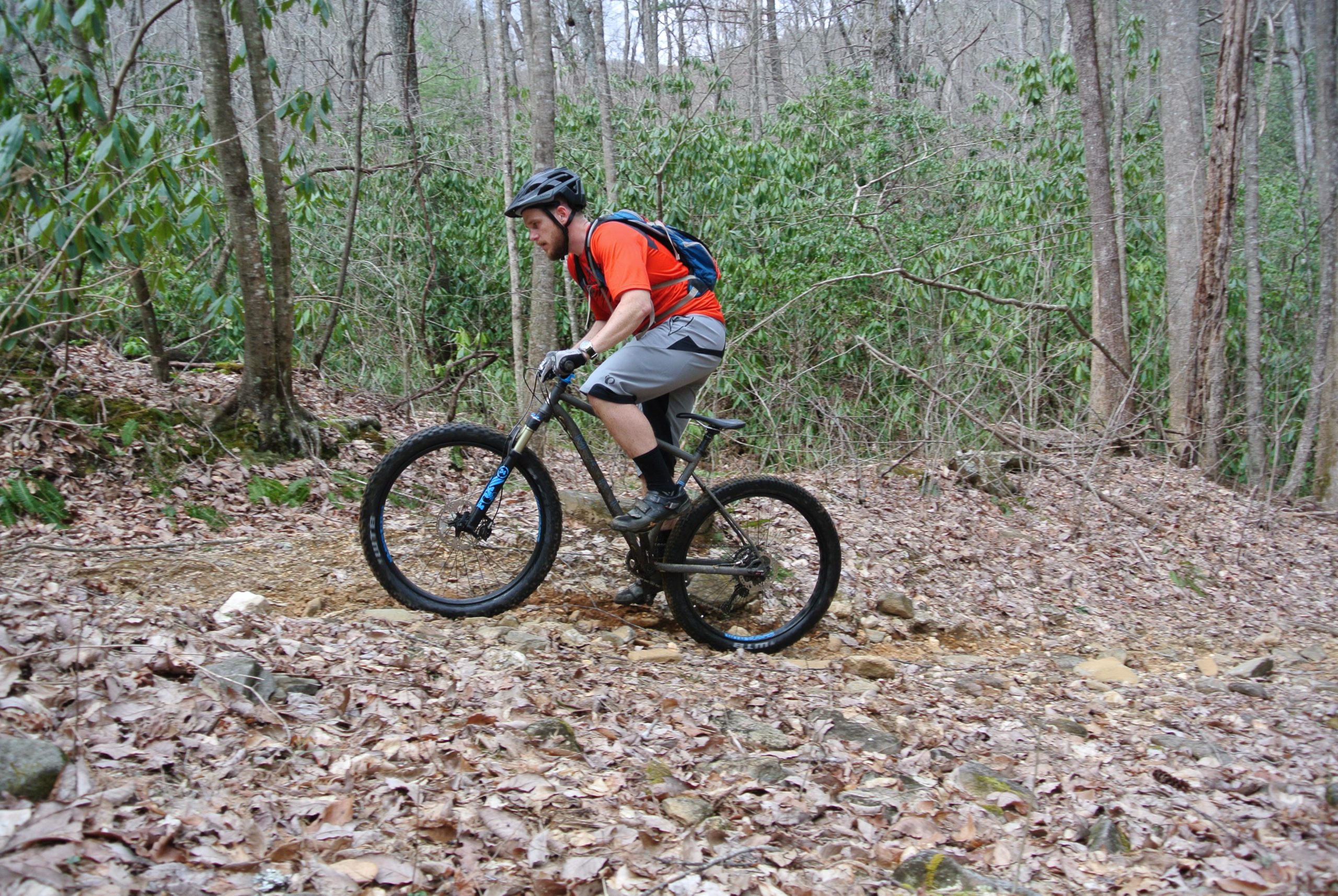 A mountain biker navigating a rugged trail surrounded by trees and underbrush, with fallen leaves and rocks on the ground. The rider, wearing an orange shirt and a helmet, is balancing on one wheel while the other wheel hovers above the terrain. Bull / Jake Mountain mountain bike trail.