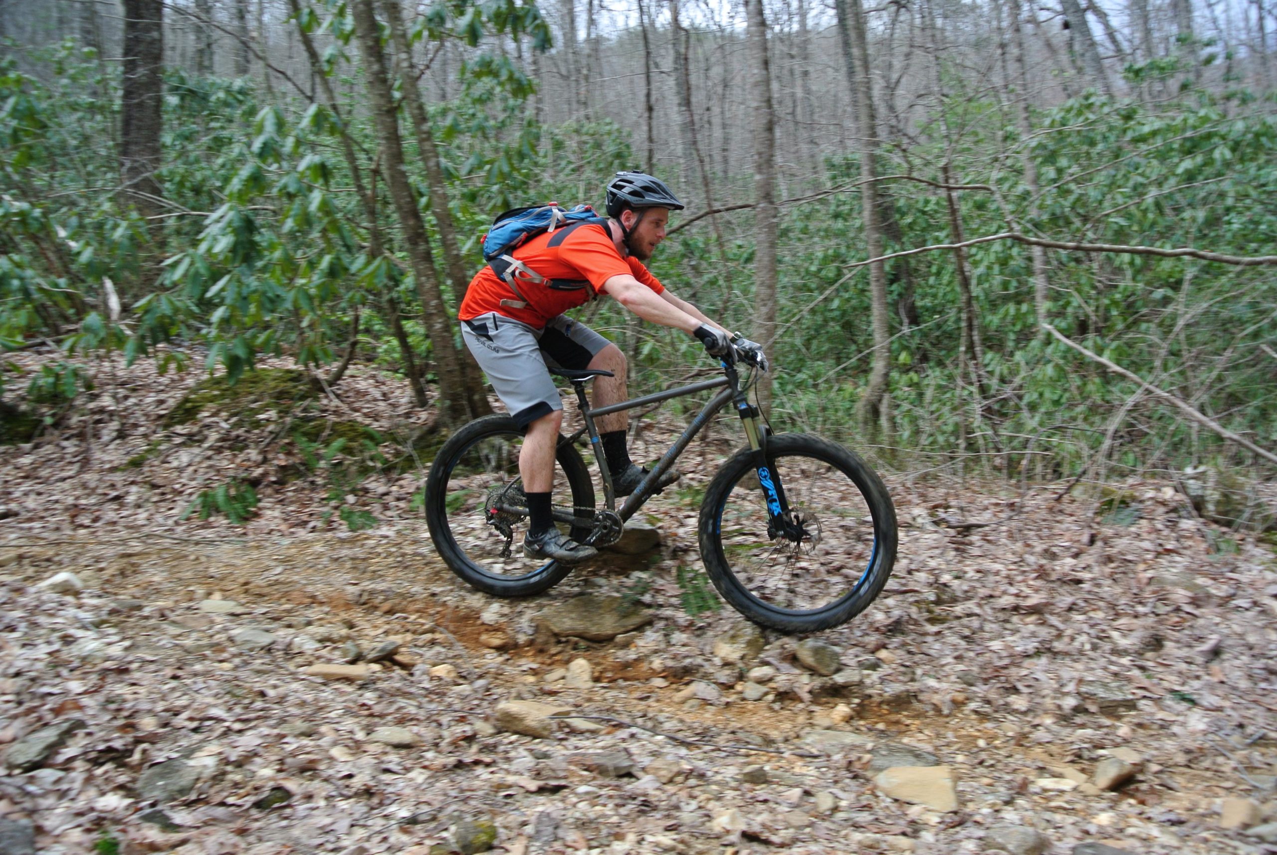 A mountain biker in an orange shirt and gray shorts jumps off a rock on a forest trail, surrounded by trees and greenery. The ground is covered with leaves and rocks, suggesting a rugged outdoor environment. The biker is wearing a helmet and has a blue backpack, focused on maintaining balance while navigating the trail. Bull / Jake Mountain mountain bike trail.