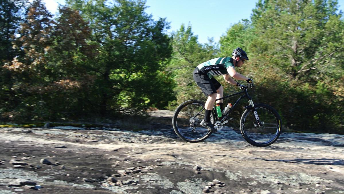 A cyclist in a green and black jersey is mid-air while performing a jump on rocky terrain, surrounded by green trees and blue skies. The rider is wearing a helmet and gloves, and a water bottle is attached to the bike frame. Georgia International Horse Park mountain bike trail.