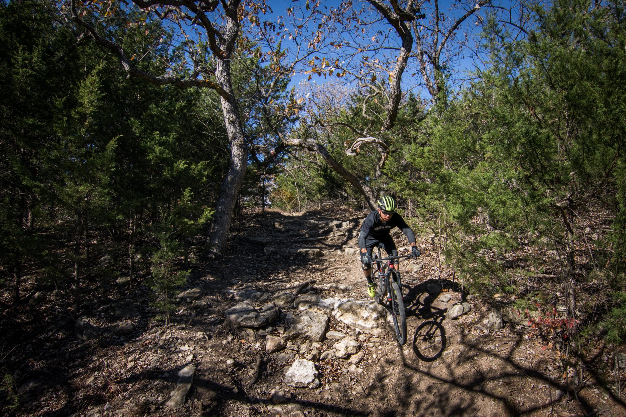 A mountain biker riding down a rocky trail surrounded by trees on a sunny day. The cyclist is wearing a helmet and dark clothing, with a focus on navigating the uneven terrain. Camp Horizon mountain bike trail.