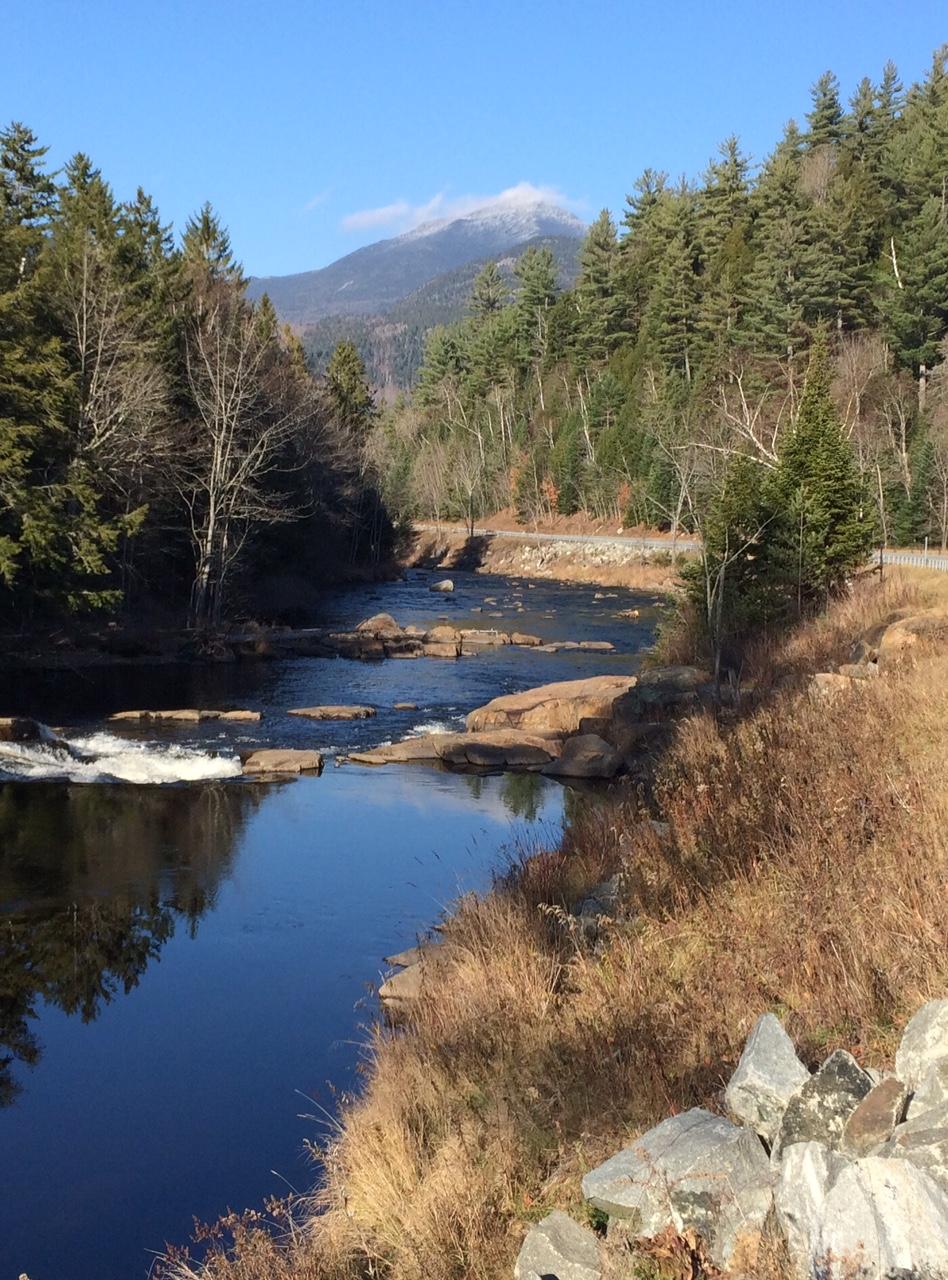 A tranquil river flows through a natural landscape, with rocky banks and trees lining both sides. In the background, a mountain is partially covered with a light dusting of snow against a clear blue sky. The scene captures the serenity of nature with reflections shimmering on the water's surface. Flume Trails mountain bike trail.