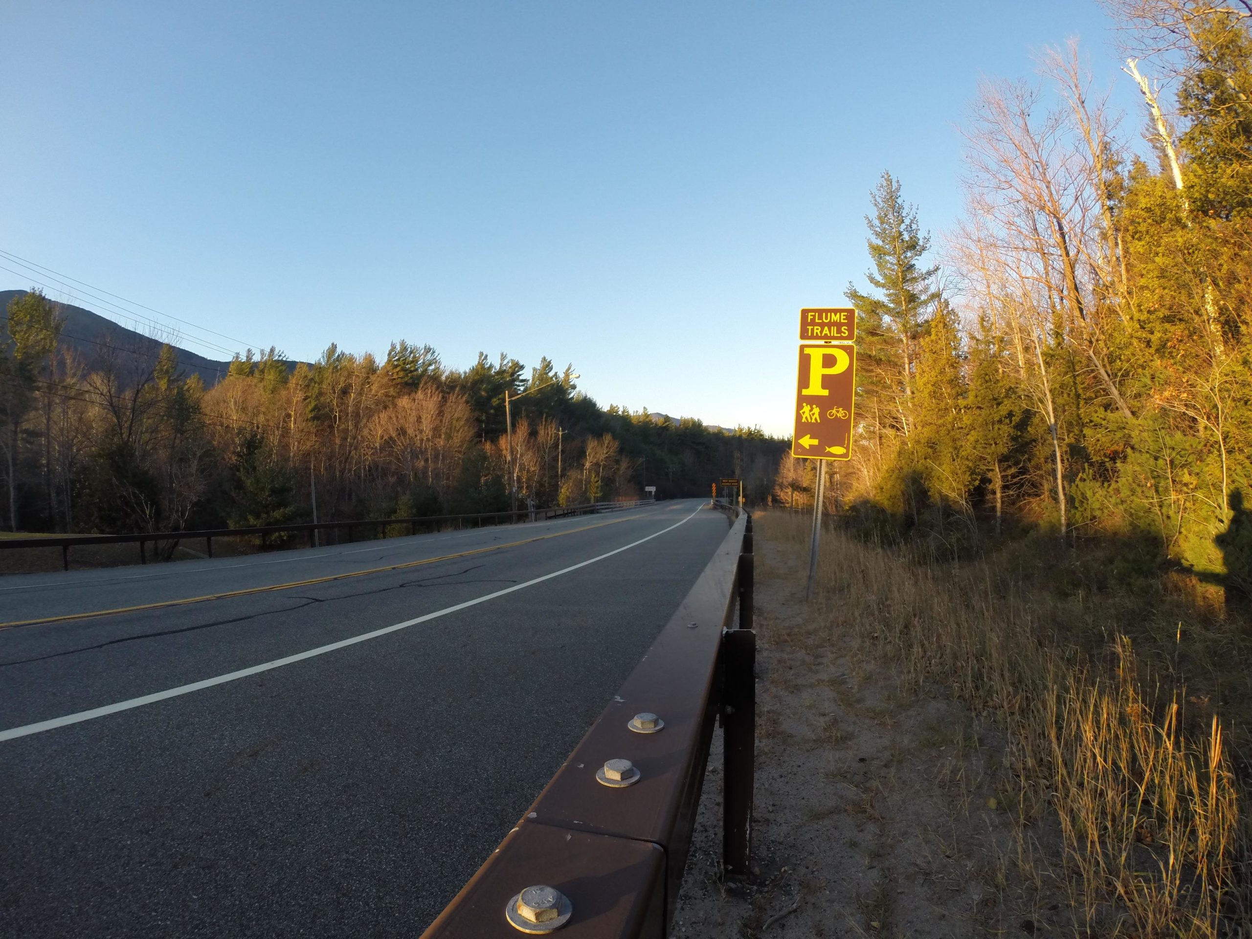 A roadside view featuring a sign for "Flume Trails" with a parking symbol, indicating areas for pedestrians and cyclists. The scene is set against a backdrop of trees and a clear sky, with a visible roadway and guardrail. The lighting suggests early morning or late afternoon. Flume Trails mountain bike trail.