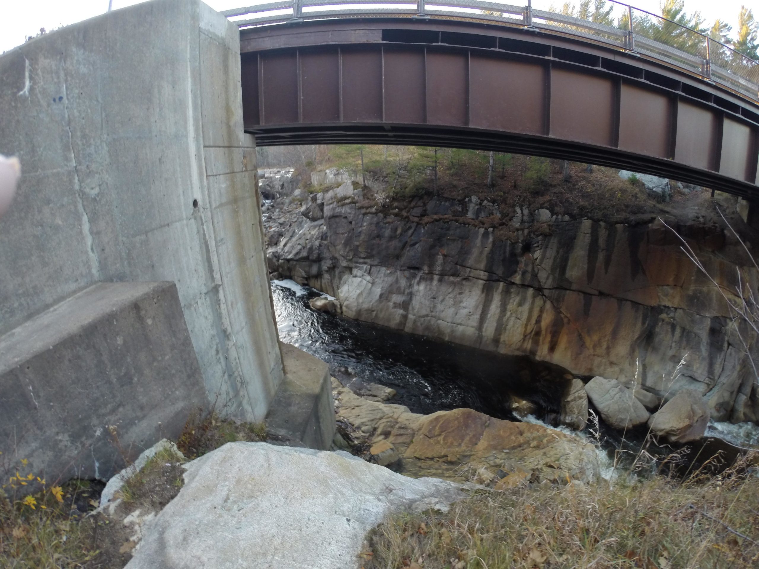 A view of a rocky riverbank with a concrete wall and a metal bridge overhead. The water flows beneath the bridge, surrounded by rocks and sparse vegetation. Flume Trails mountain bike trail.