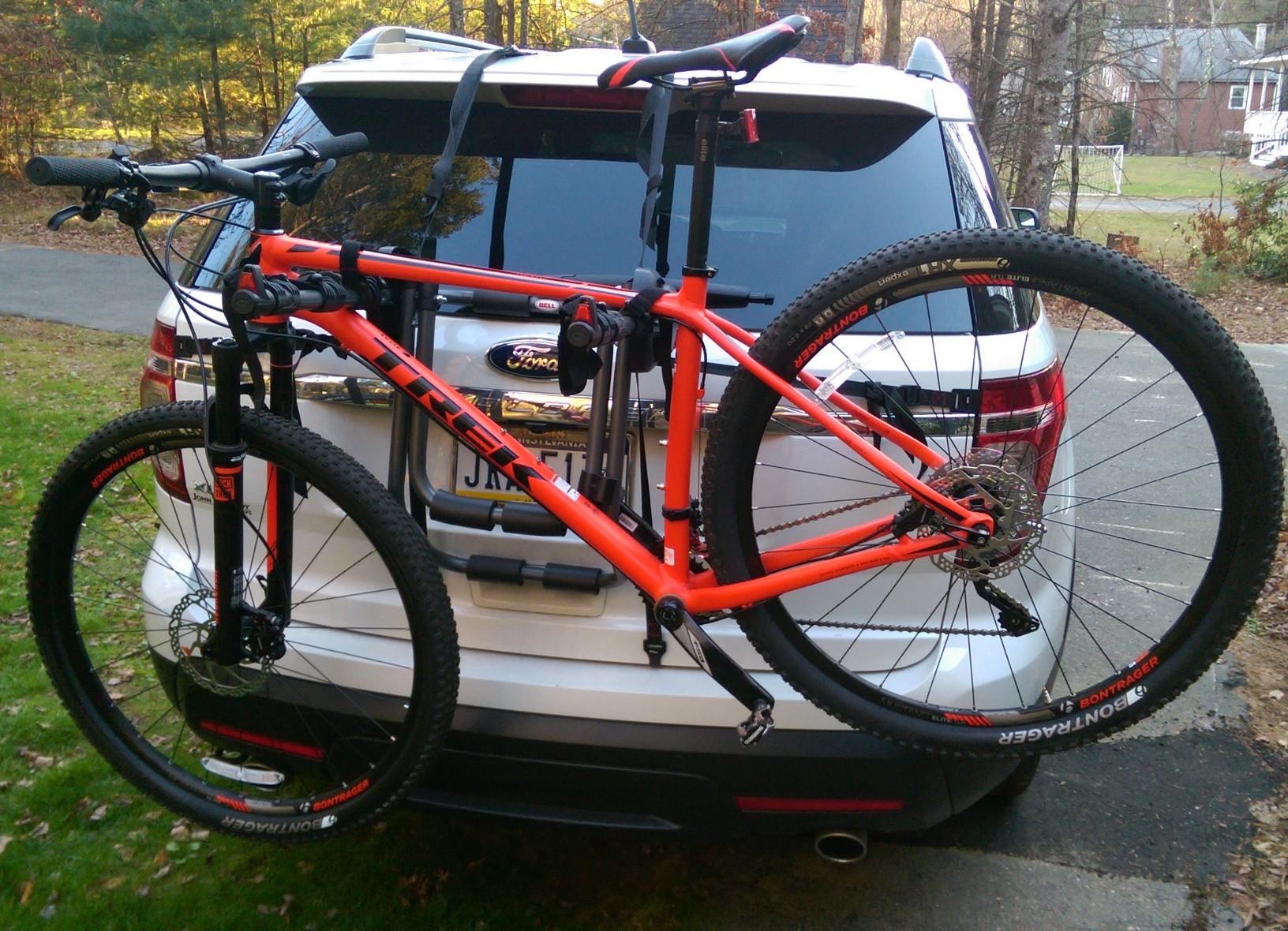 Trek Superfly 7: A bright orange mountain bike mounted on the back of an SUV. The bike is securely attached with straps and features black tires. The vehicle is parked in a grassy area, with trees visible in the background.