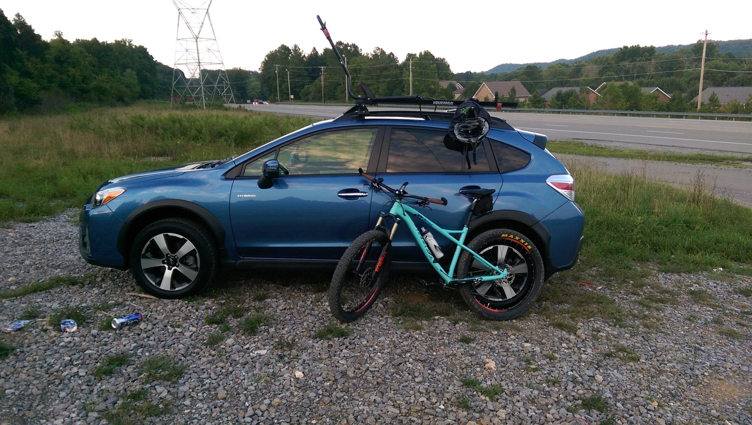 Orbea Loki: A blue hybrid SUV parked on a gravel area beside a road. A mountain bike with a turquoise frame is leaning against the vehicle, and a helmet is attached to the car's roof rack. In the background, there are grass and trees, along with a power line tower visible in the distance.