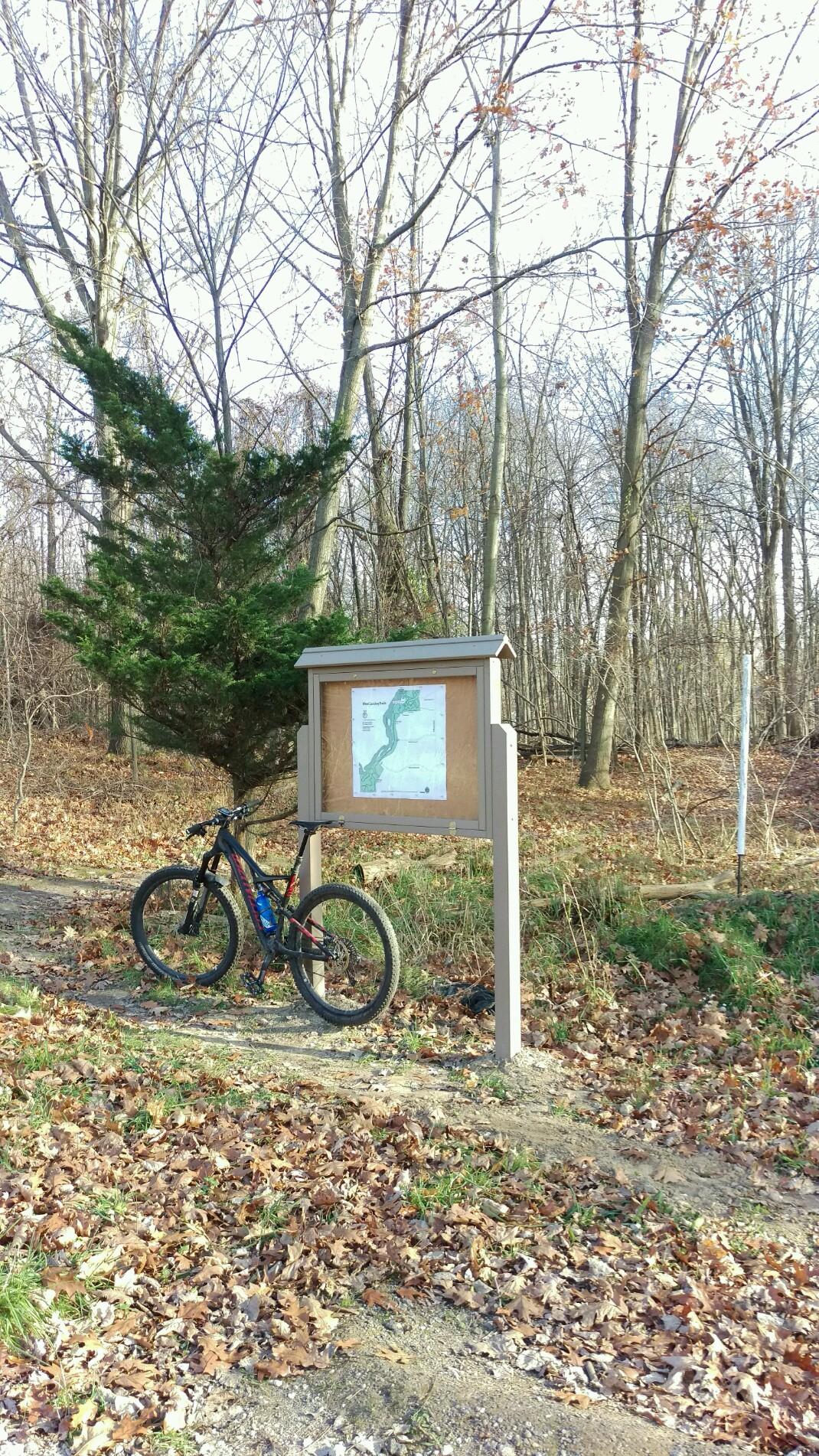 A mountain bike parked beside a trail map sign in a wooded area with bare trees and fallen leaves covering the ground. Hunters Ridge mountain bike trail.