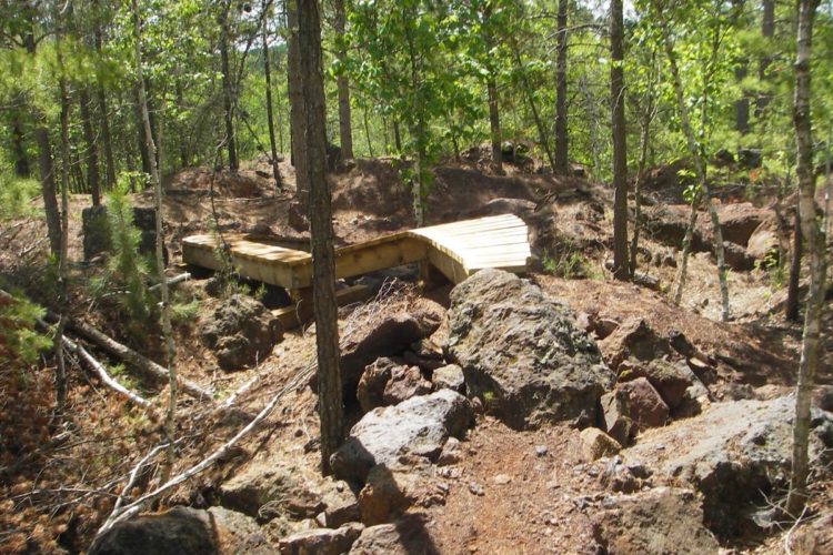 A wooden bridge spans a rocky terrain, surrounded by lush green trees and underbrush in a forest setting. The area features uneven ground with scattered rocks and fallen branches.