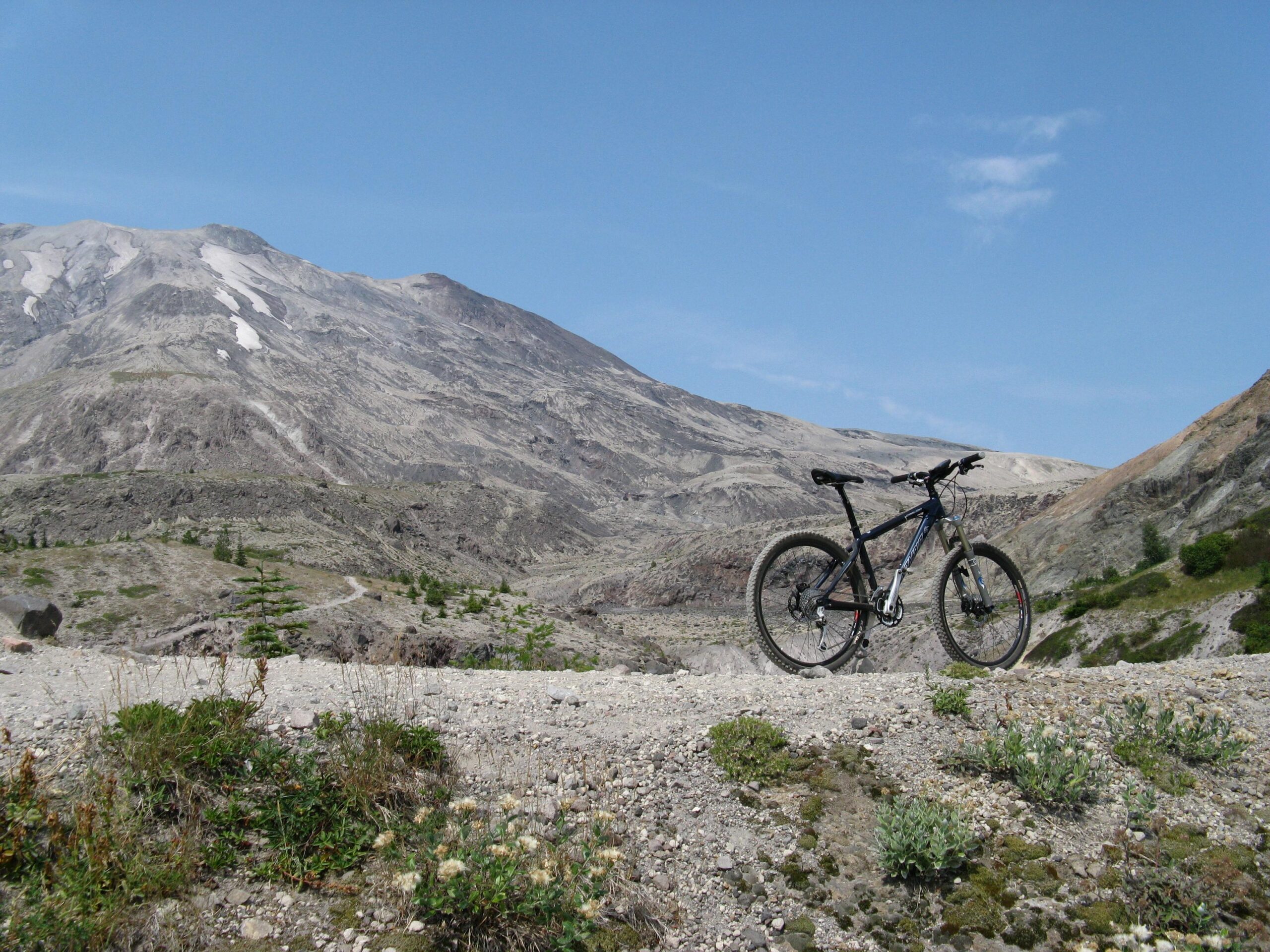 Santa Cruz Chameleon: Mountain bike standing on rocky terrain with a backdrop of a vast, mountainous landscape under a clear blue sky. Green patches of vegetation are visible in the foreground, contrasting with the gray tones of the terrain.