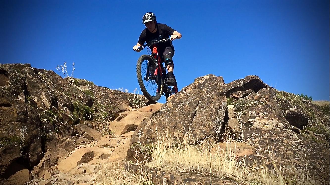 A mountain biker navigating rocky terrain against a bright blue sky, lifting the front wheel of the bike as they prepare to descend over the rocks. Sparse vegetation and dry grass surround the path. Syncline mountain bike trail.