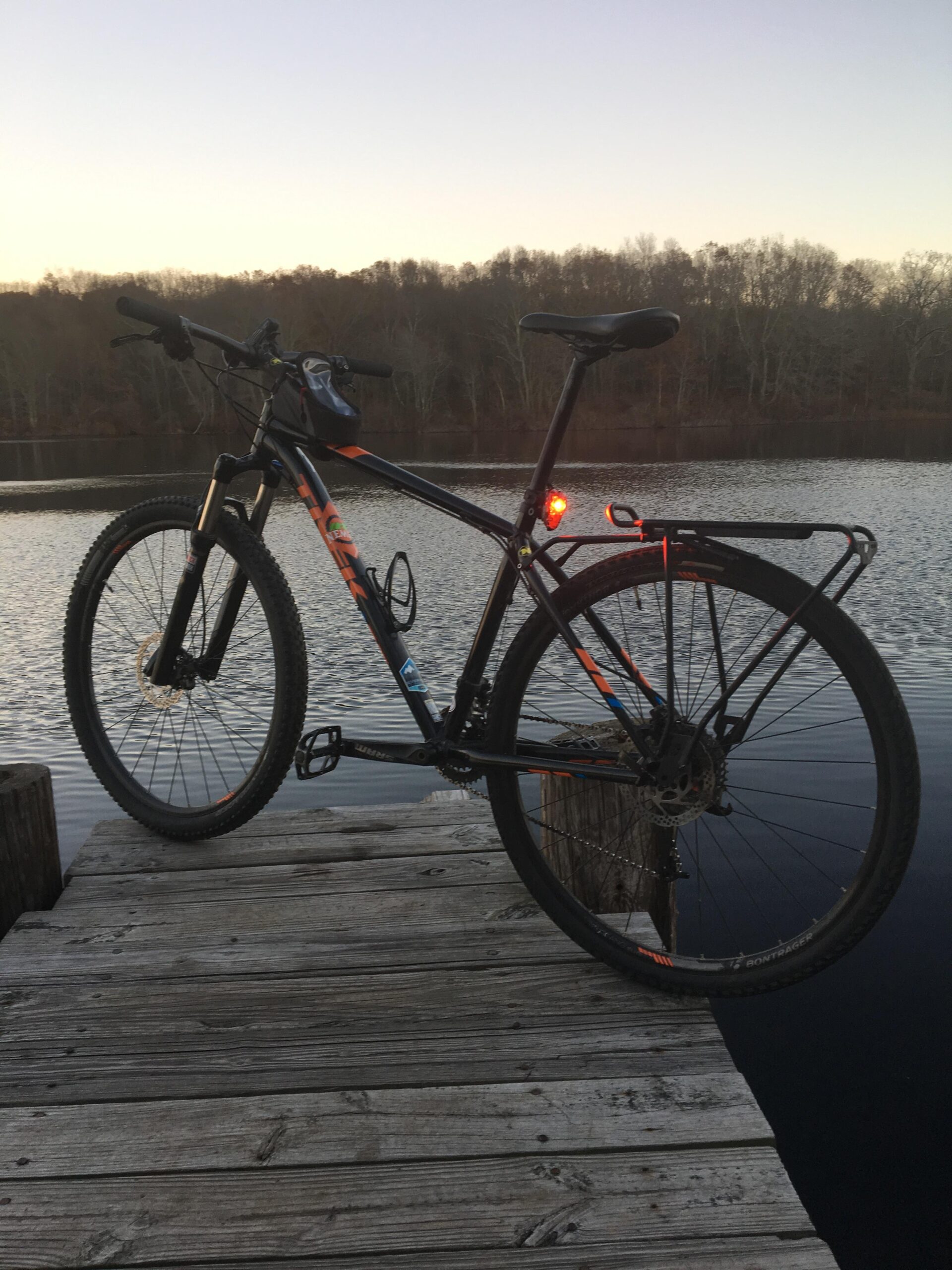 Trek X-Caliber 8: A mountain bike is parked on a wooden dock overlooking a calm body of water at sunset, with trees in the background. The bike features a rear light and a small bag attached to the handlebars.