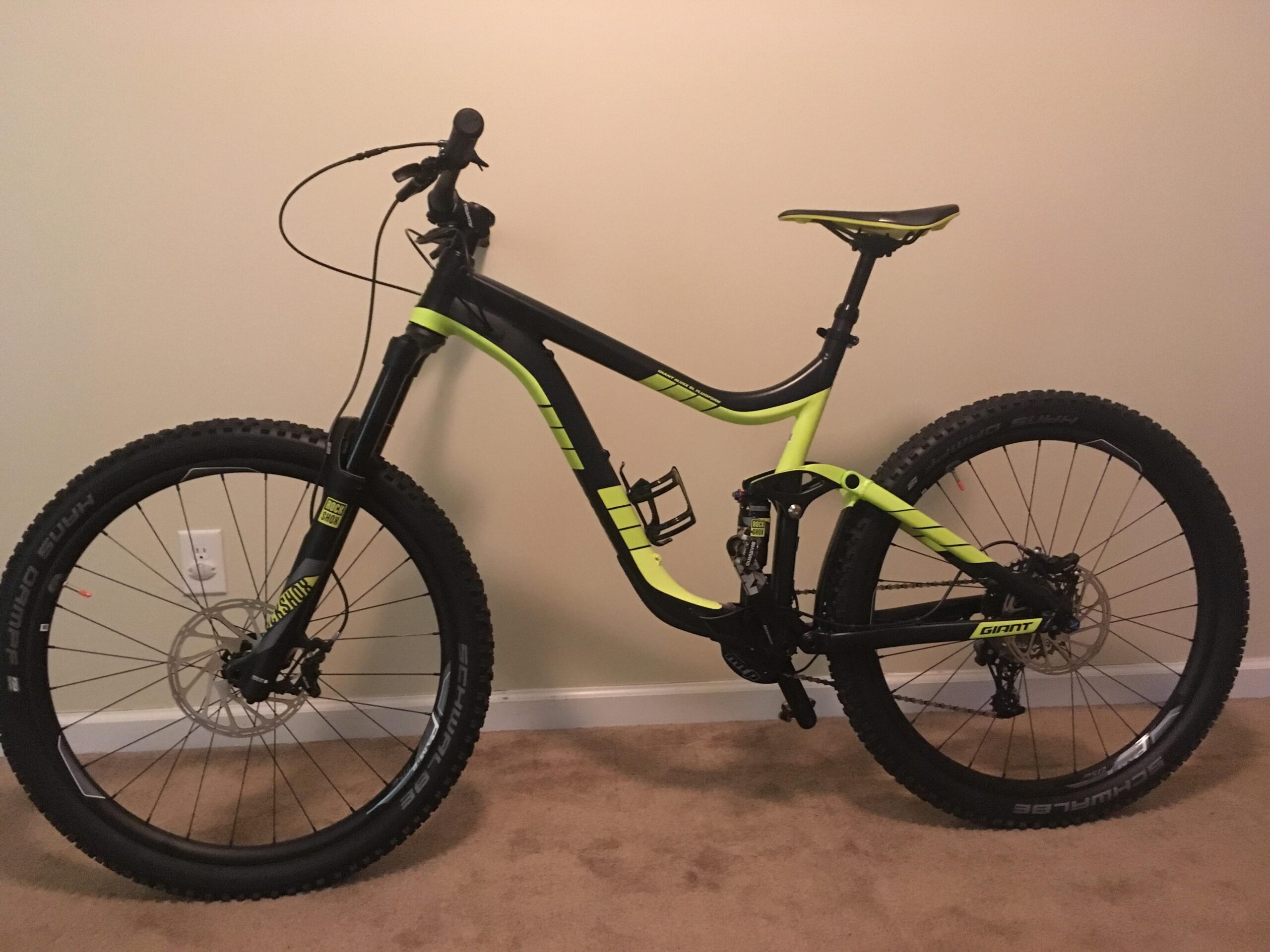 Giant Reign: A black and neon yellow mountain bike leaning against a wall, showcasing its large tires, dual suspension, and modern design features. The bike's frame displays a sleek, sporty appearance, and it is equipped with a disc brake and a gear-shifting mechanism, positioned on a carpeted floor.