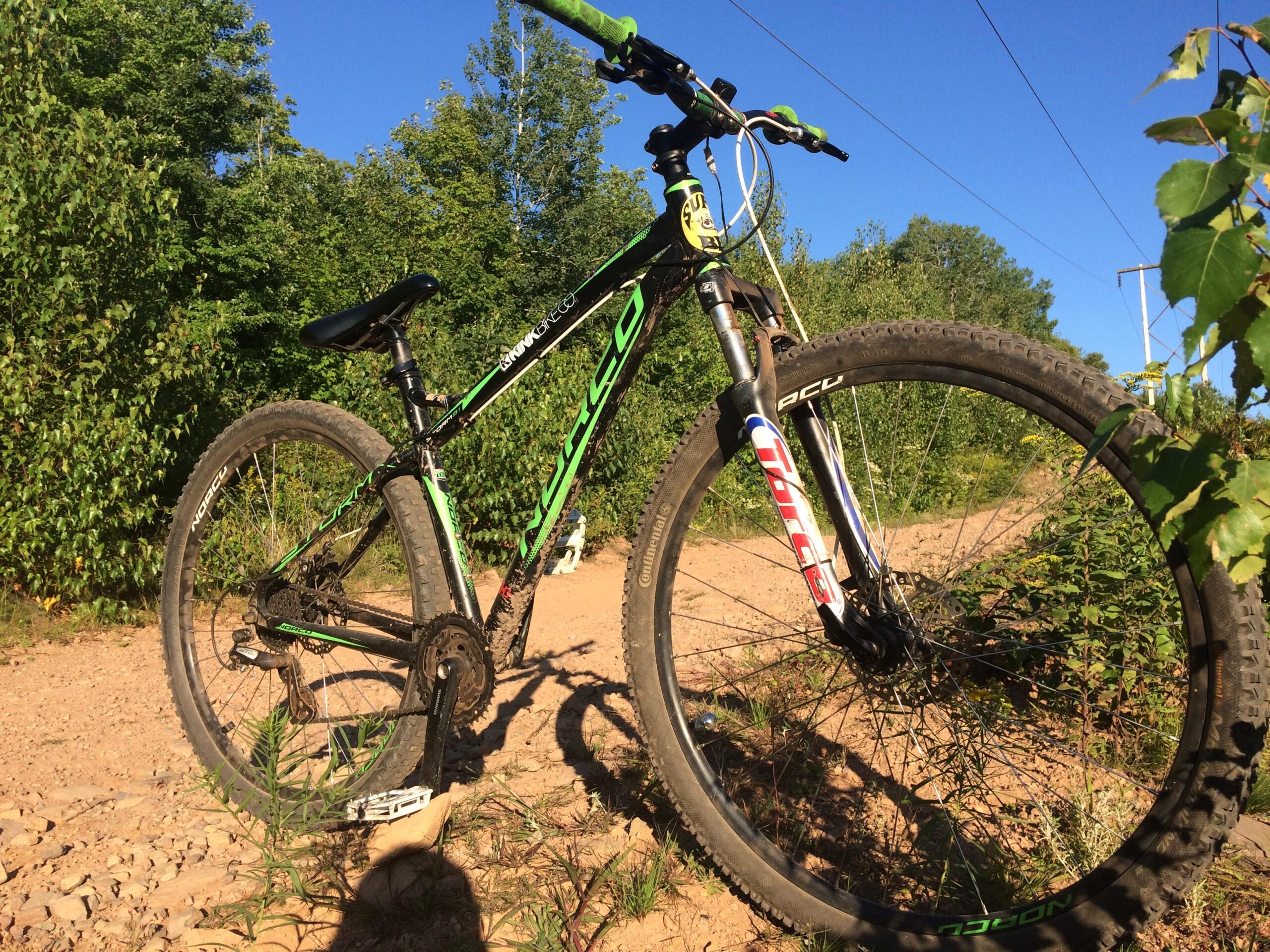 Norco Storm: A close-up view of a green and black mountain bike resting on a gravel trail, surrounded by lush greenery under a clear blue sky. The bike features thick tires, a sturdy frame, and visible gear components.