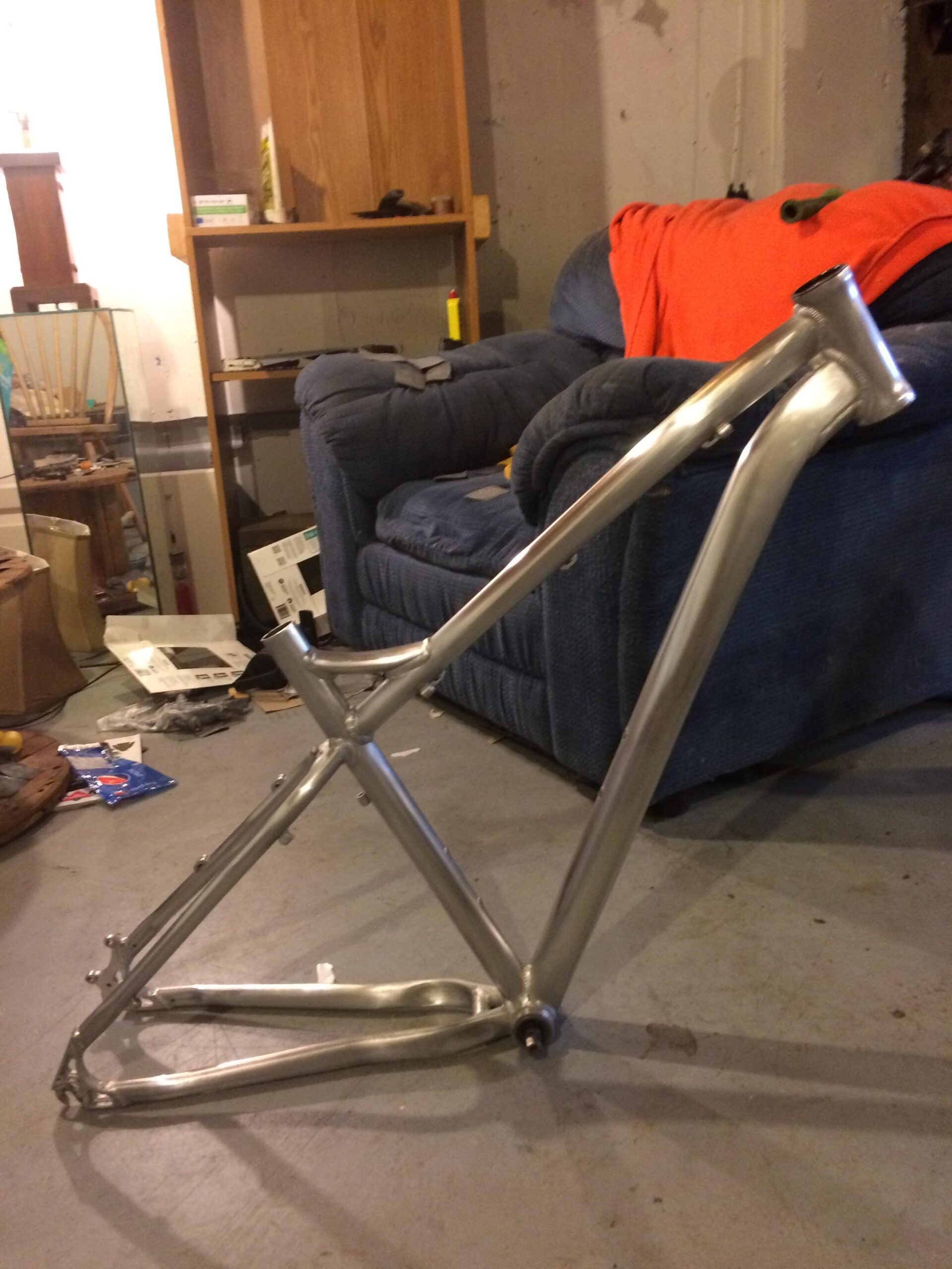 Norco Storm: A metallic bicycle frame leaning against a blue couch in a cluttered basement setting. The background includes shelves with items, a couch covered with an orange blanket, and various objects scattered on the floor.