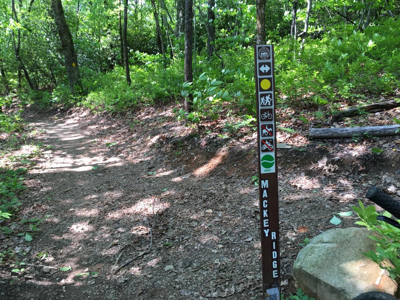 A dirt trail winding through a lush forest with a signpost marking the Mackey Ridge trail. The sign features various symbols indicating the activities allowed on the trail, including hiking, biking, and wildlife viewing. Sunlight filters through the trees, illuminating the pathway and surrounding greenery. Bracken Preserve mountain bike trail.