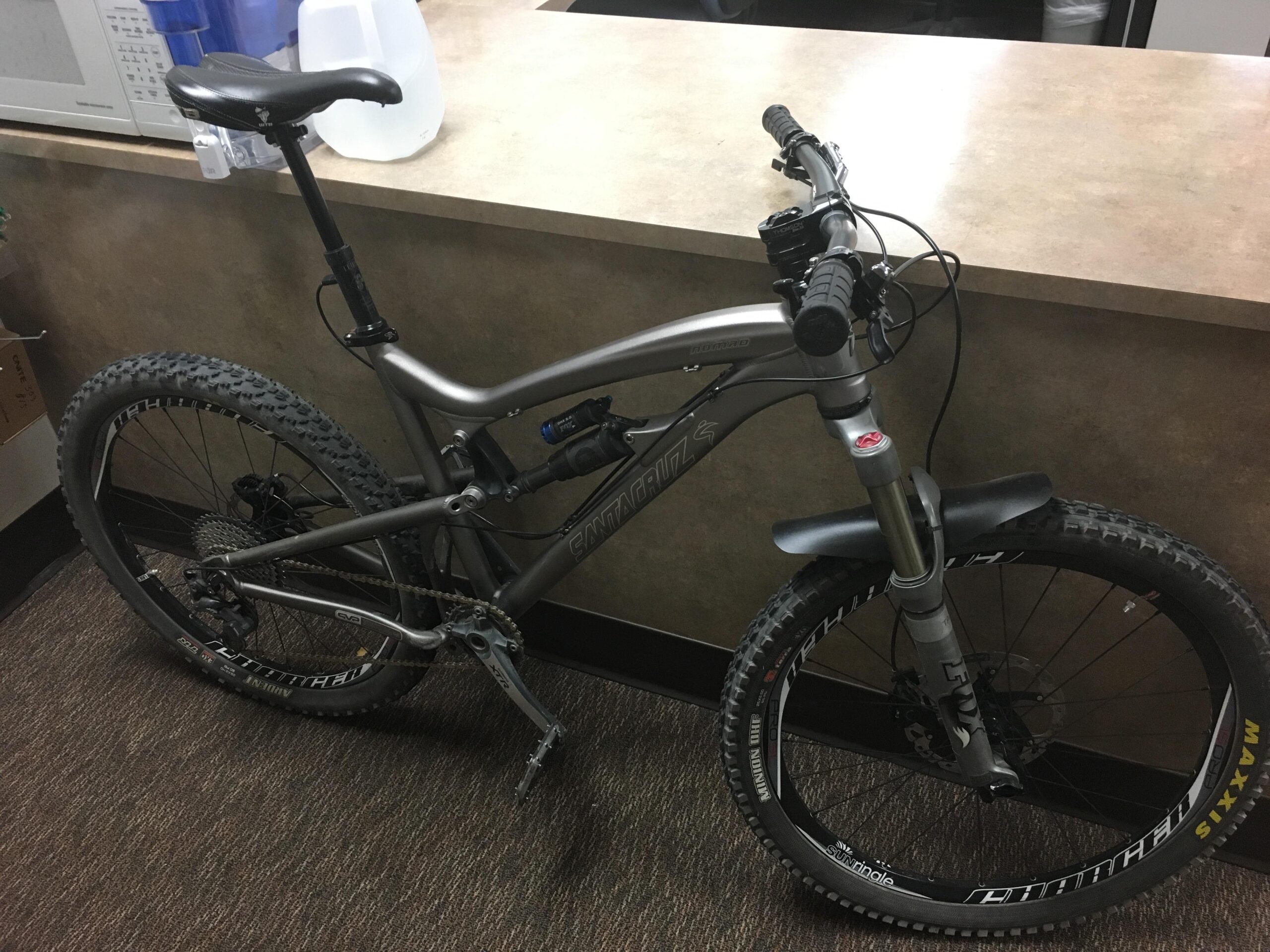 Santa Cruz Nomad: A mountain bike with a gray frame is leaning against a counter. The bike features thick tires suitable for off-road riding, a suspension fork, and a dropper seat post. A jug of water is visible on the counter beside the bike. The setting appears to be indoors, with a neutral-colored background.