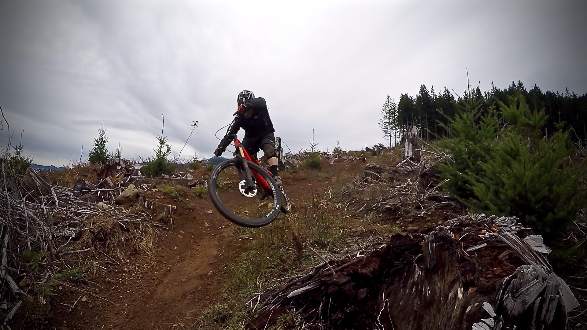 A mountain biker in a black outfit jumps over a rugged terrain on a red bicycle. The background features a cloudy sky, scattered tree stumps, and young conifer trees, creating a natural forest setting. Cold Creek mountain bike trail.