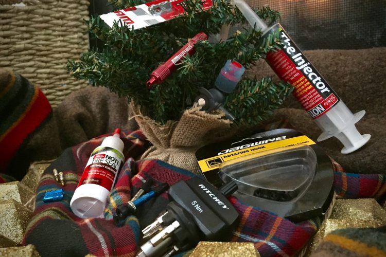 A small artificial Christmas tree surrounded by various bike maintenance tools and products, including a bottle of tire sealant, a syringe labeled "The Injector," and a rotating hook. The items are placed on a plaid cloth with decorative golden elements, and there is a warm background with soft lighting.