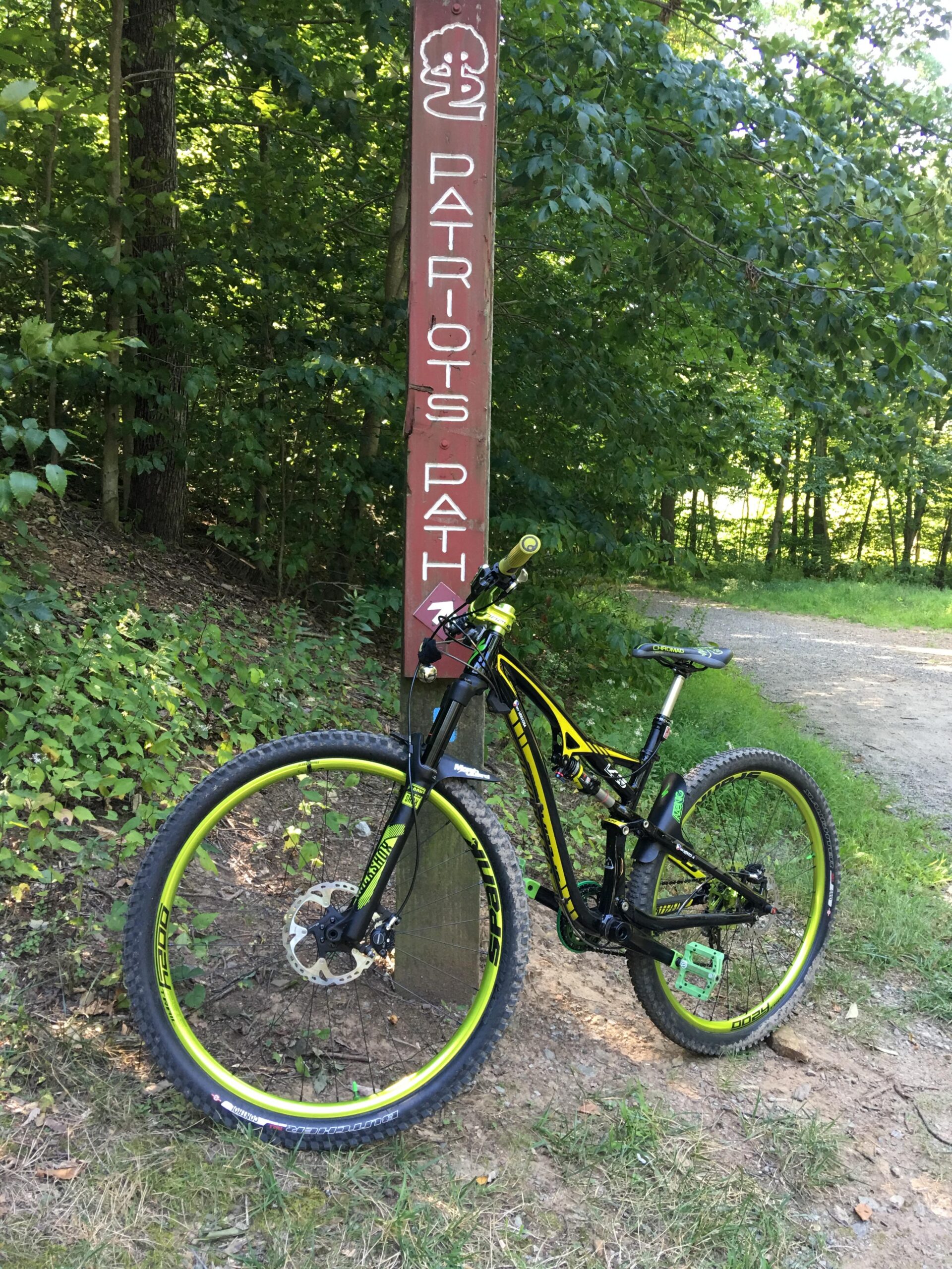 Specialized Camber Comp 29: A black and yellow mountain bike is leaning against a wooden trail sign that reads "Patriots Path." The background is a lush green forest with trees and foliage. The path is gravel, indicating a biking or hiking trail.