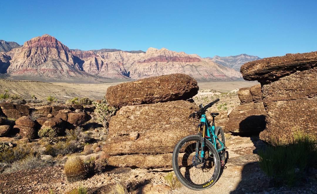 A mountain bike rests on a rocky trail, framed by large boulders in a desert landscape. In the background, vibrant red and orange mountains contrast against a clear blue sky, showcasing the natural beauty of the area. Various desert plants and shrubs are scattered around, adding to the rugged scenery. Cowboy Trails mountain bike trail.