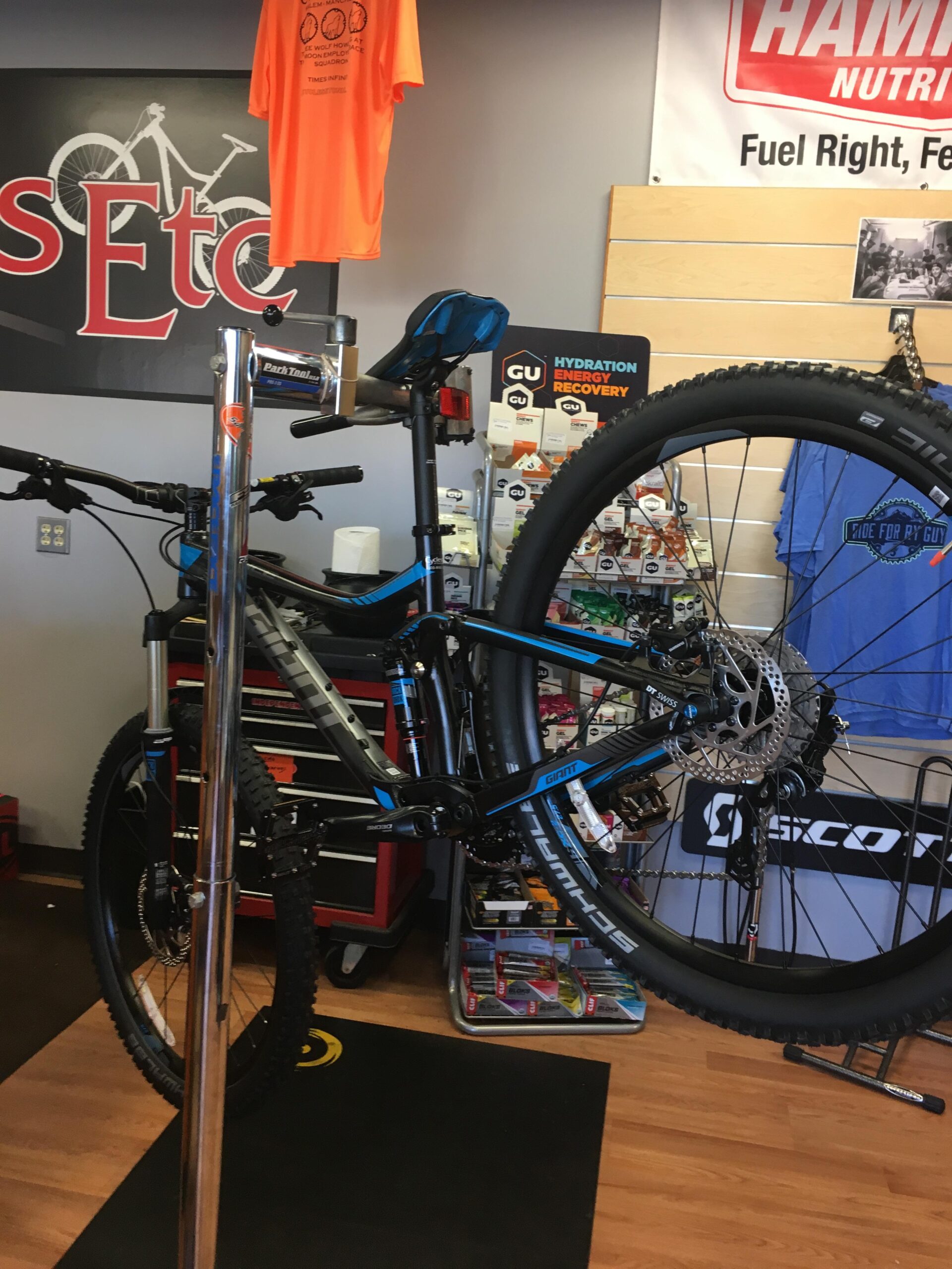 Giant trance 3: Mountain bike on a repair stand in a bike shop, displaying various accessories and nutrition products in the background. An orange t-shirt is hanging on the wall, and there are shelves with cycling-related items. The shop has a wooden floor and a logo visible on the wall.