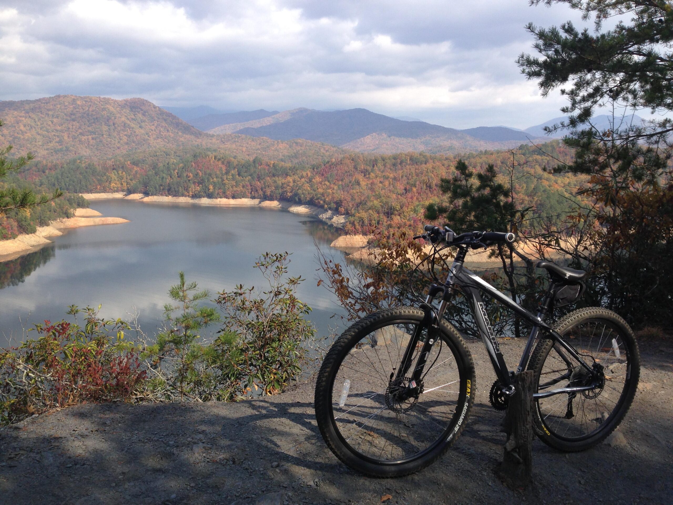 Marin Alpine Trail 29er: A mountain bike rests on a rocky outcrop with a scenic view of a lake surrounded by colorful autumn foliage and distant mountains under a partly cloudy sky.