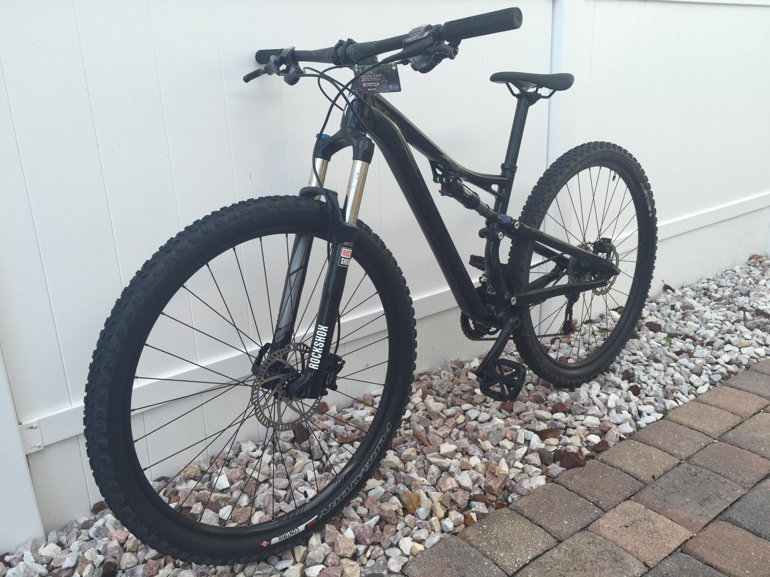 Specialized Camber 29: A black mountain bike leaned against a white fence, positioned on a bed of small stones. The bike features thick, knobby tires suitable for off-road terrain, a suspension fork, and a modern design. The area below the bike consists of arranged paving stones.