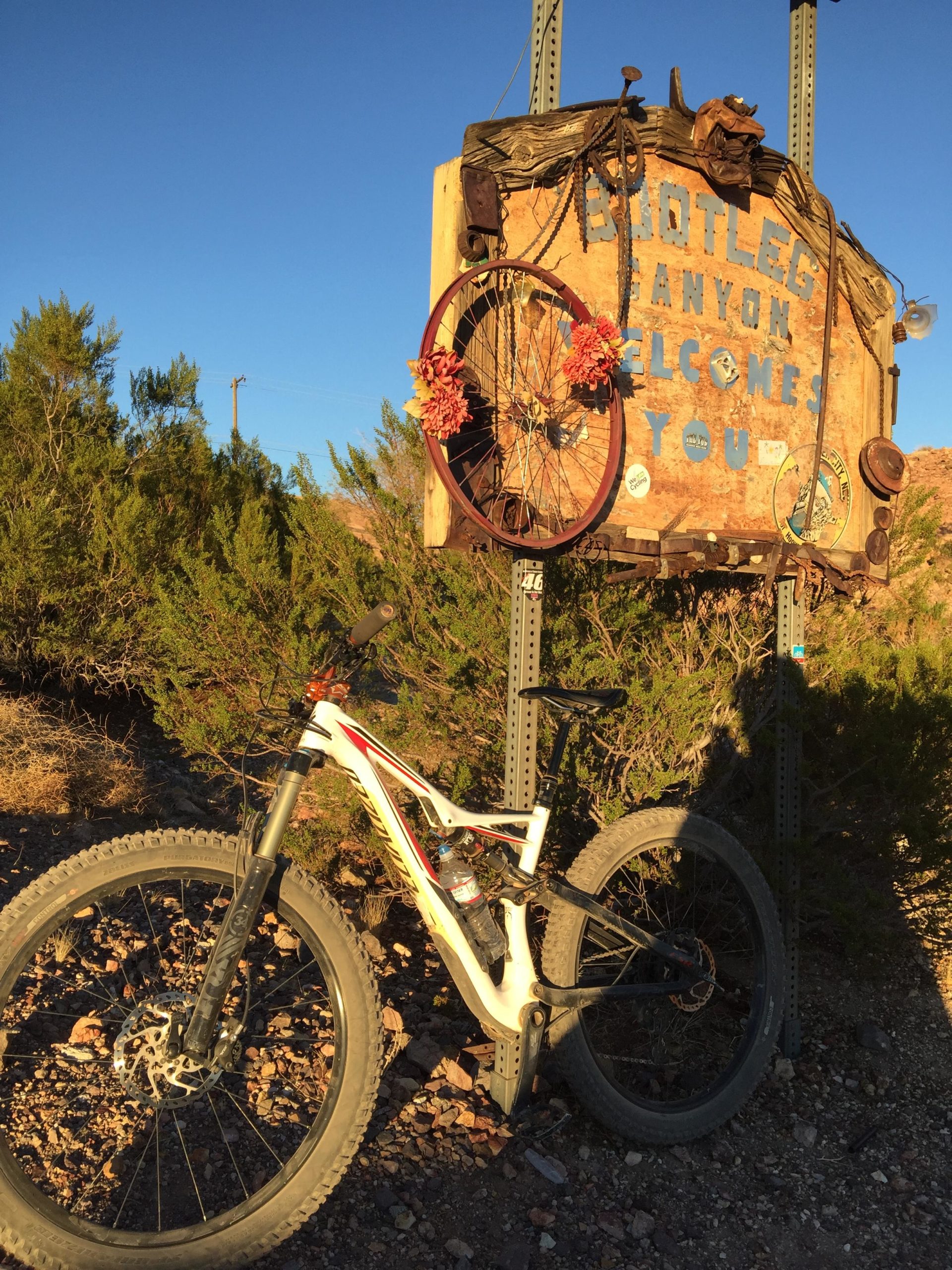 A mountain bike leans against a weathered wooden sign that reads "Bootleg Canyon Welcomes You," adorned with a vintage bicycle wheel and flowers. The sign is set against a clear blue sky and surrounded by desert foliage. Bootleg Canyon mountain bike trail.