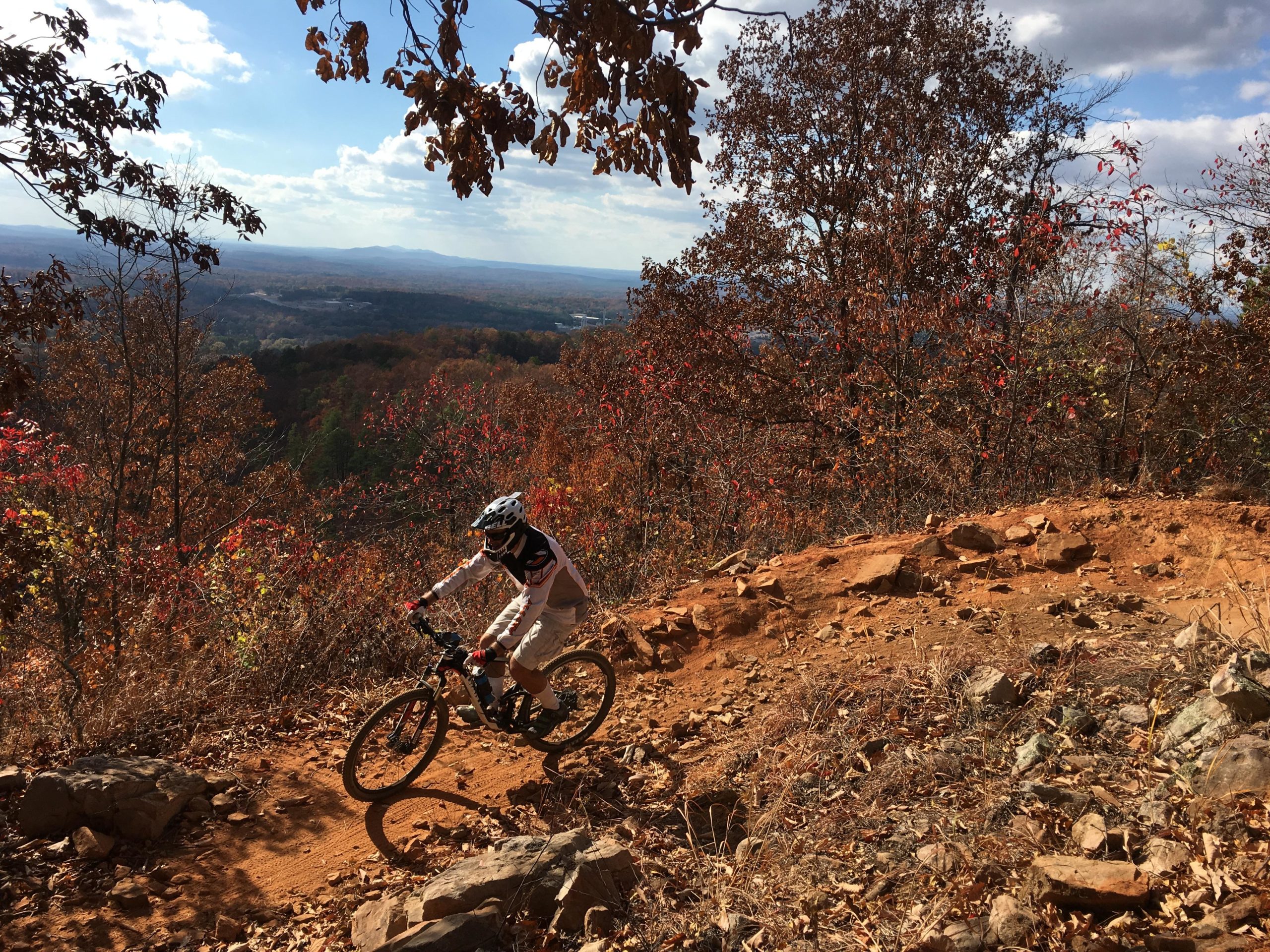 A mountain biker navigating a rocky trail with autumn foliage in the background, set against a vibrant landscape of hills and a cloudy sky. Coldwater Mountain mountain bike trail.