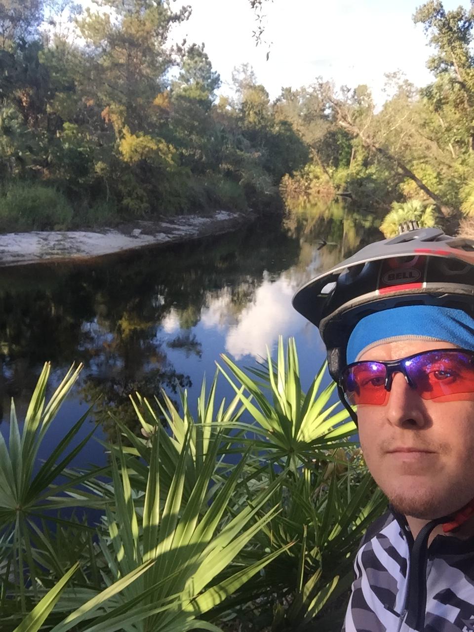 Specialized Camber 29: A person wearing a cycling helmet and sunglasses takes a selfie by a calm, reflective river surrounded by lush greenery, including palm plants and trees. The scene is bathed in soft sunlight, highlighting the natural beauty of the landscape.