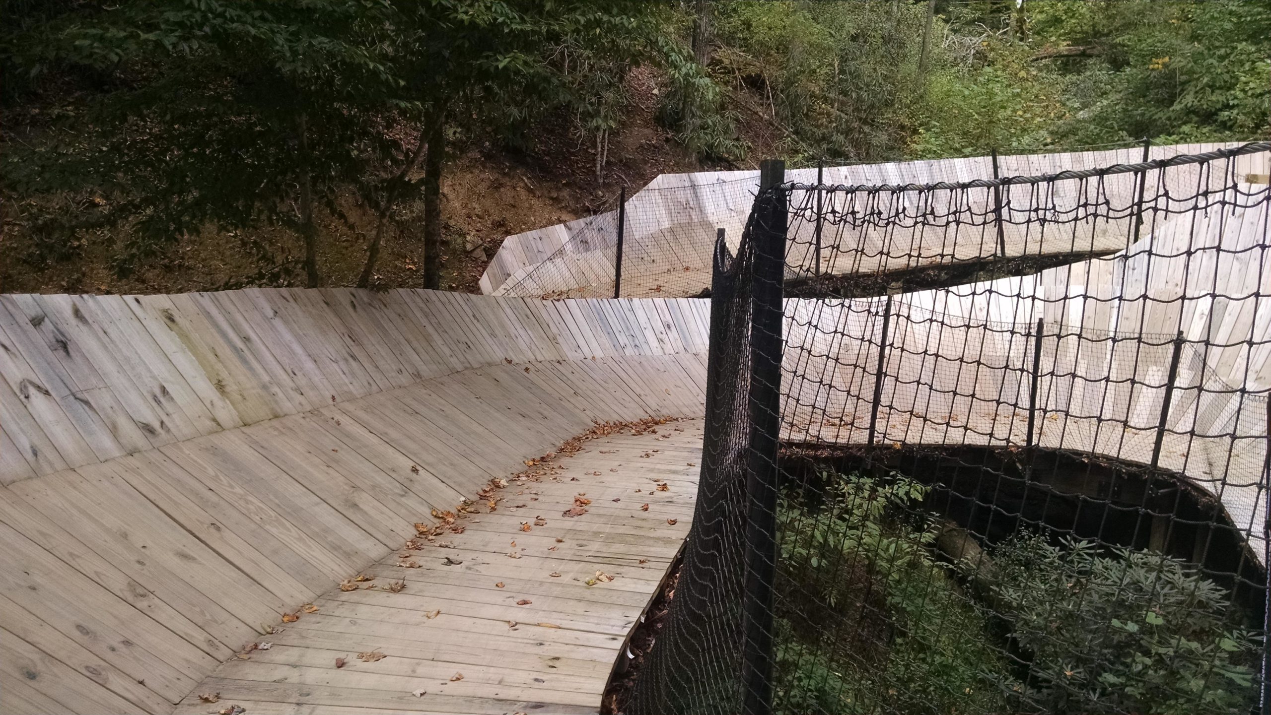 A winding wooden path through a wooded area, featuring smooth curves and a safety net on one side. Leaves are scattered on the ground, suggesting that it is autumn. The surrounding trees provide a natural backdrop, enhancing the scenic view of the path. Climbworks mountain bike trail.