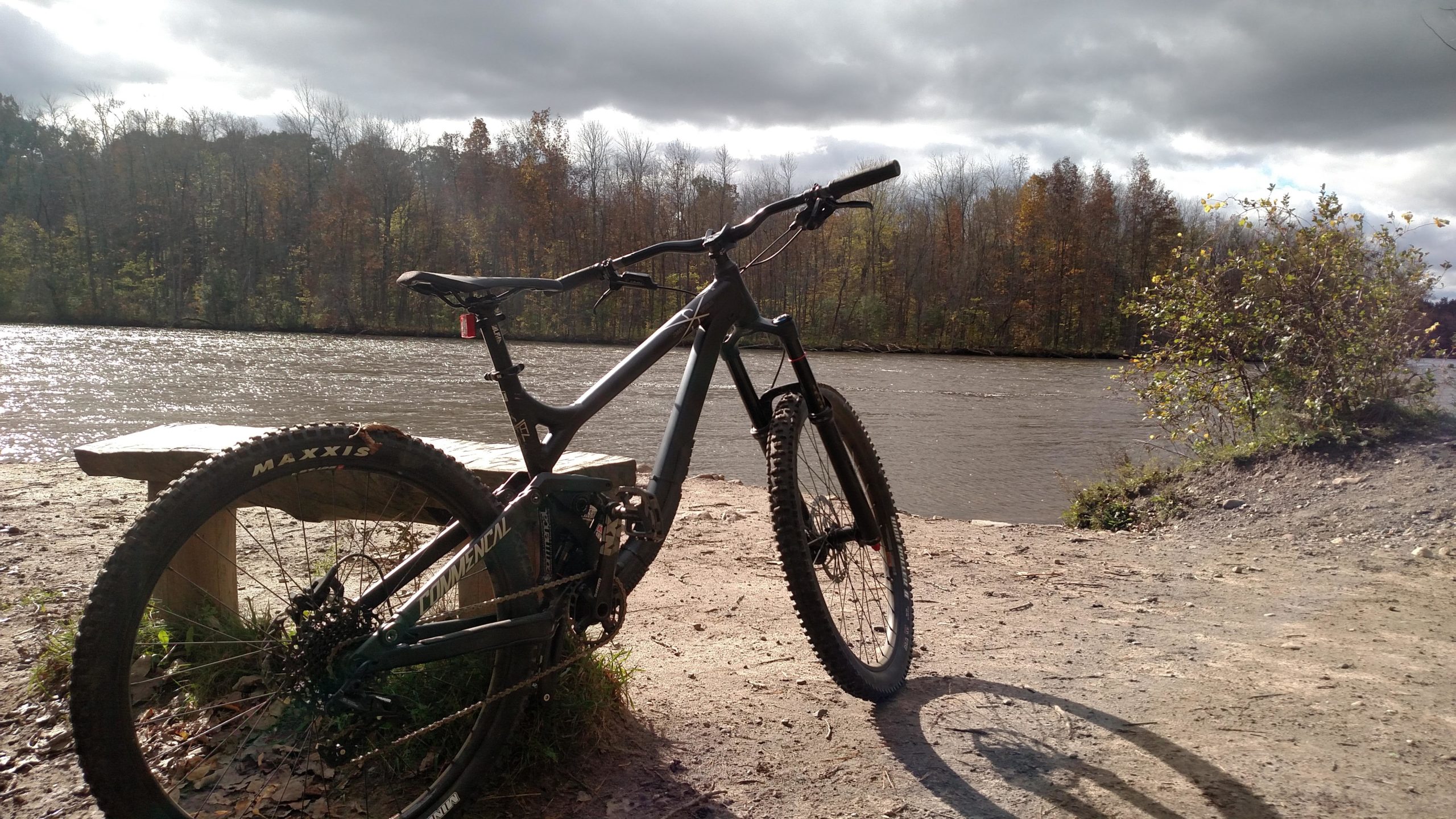 A mountain bike leaning against a wooden bench near a river, surrounded by autumn foliage and under a partly cloudy sky. Great Bear mountain bike trail.